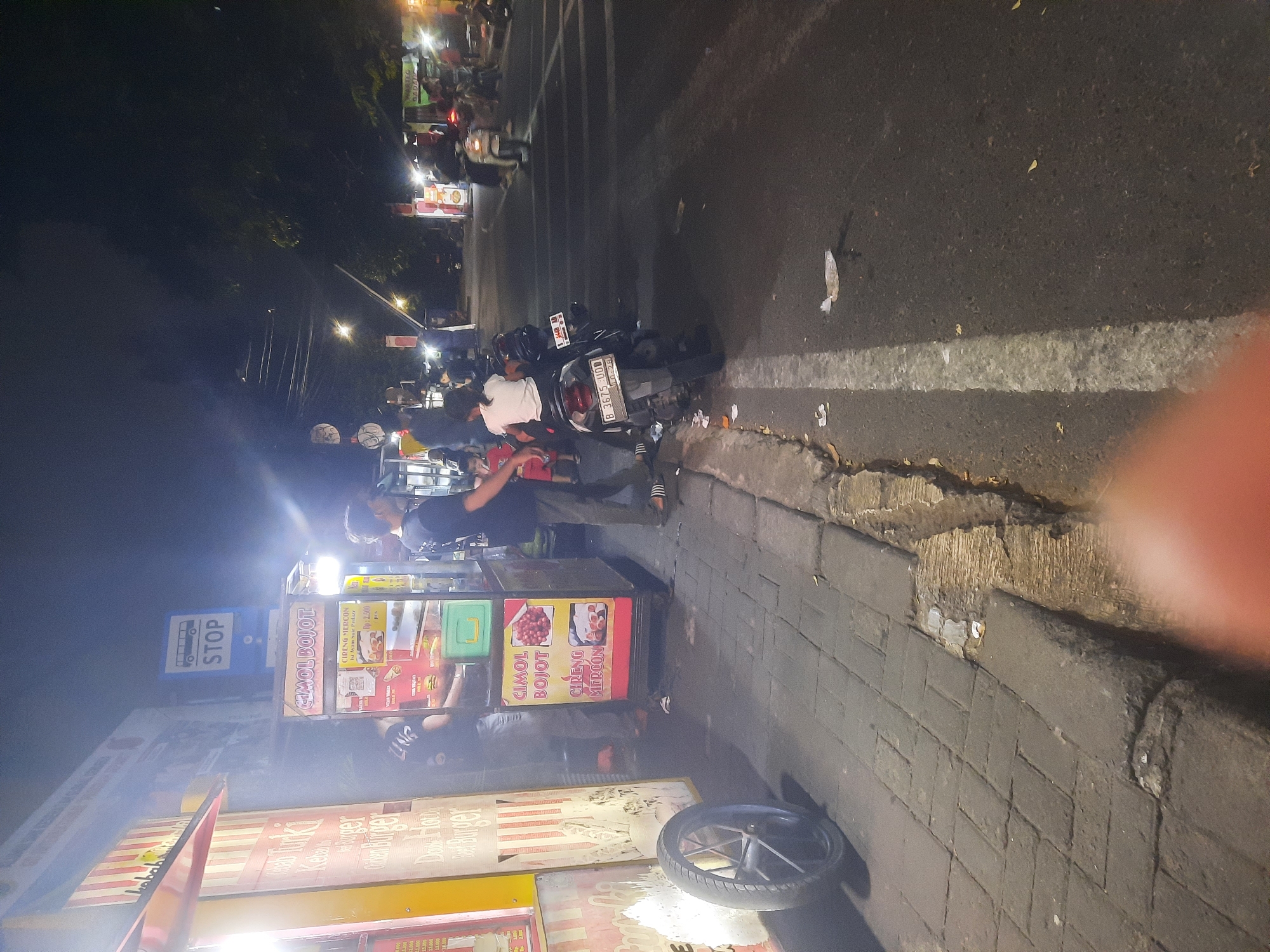 This is a nighttime street scene in North Jakarta, Indonesia, featuring a food stall with various items displayed. A man in a black t-shirt stands behind the stall, and another person in a white t-shirt is seated on a motorcycle in front of it. Several other individuals are visible in the background, some on motorcycles, suggesting a busy street. The road is paved, and there is a sidewalk with a broken curb. The overall lighting suggests it is after dark, with artificial lights from the stalls and streetlights illuminating the area.