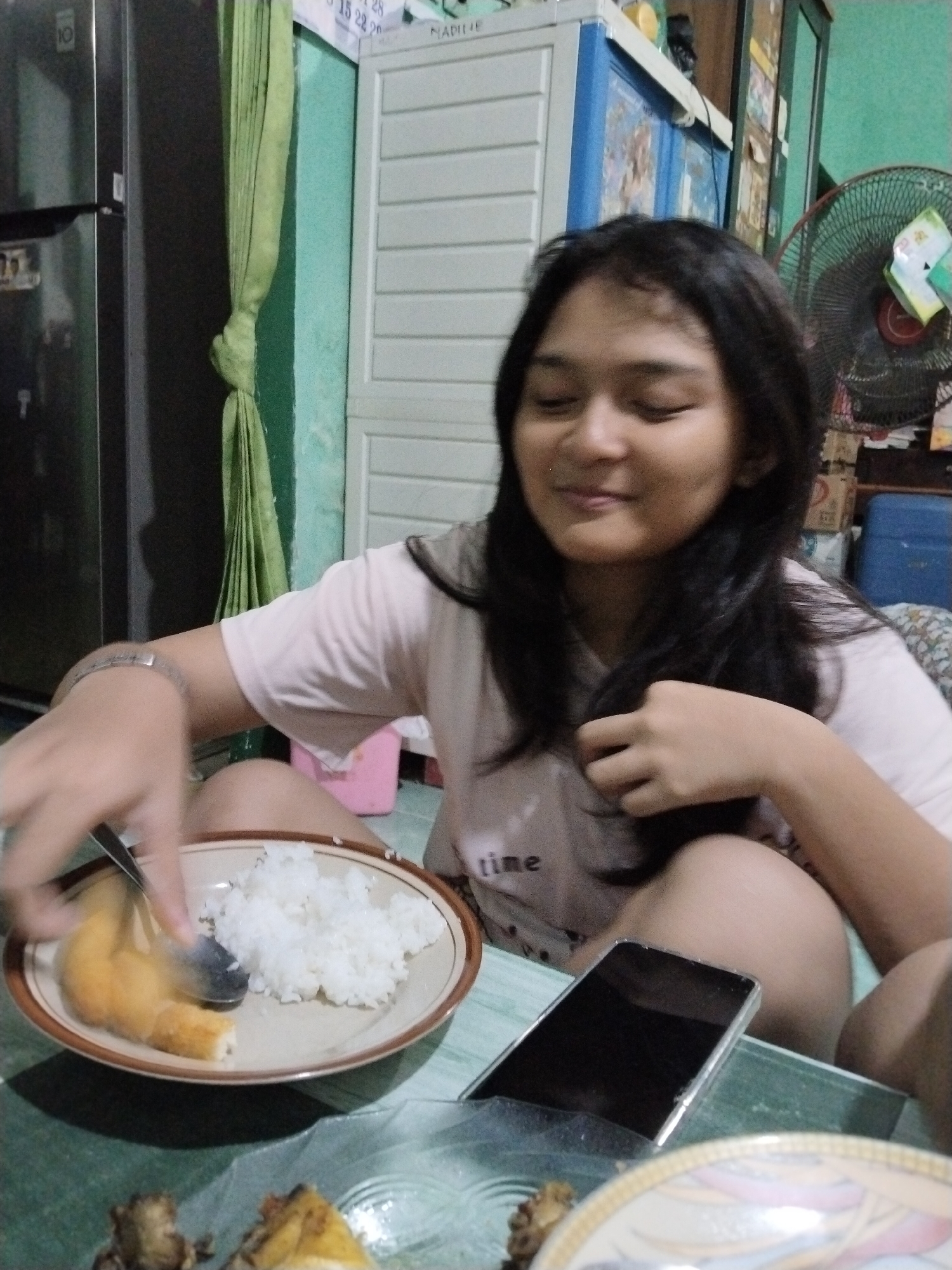 A young woman with long, dark hair is seated in a room. She is wearing a light-colored t-shirt with "time" printed on it. In front of her is a plate of white rice and a piece of fried food, which she is cutting with a spoon. A smartphone lies on the table next to the plate. In the background, there is a refrigerator, a green curtain, a white cabinet, and a fan. The overall environment suggests a residential setting in Kecamatan Pati, Indonesia.