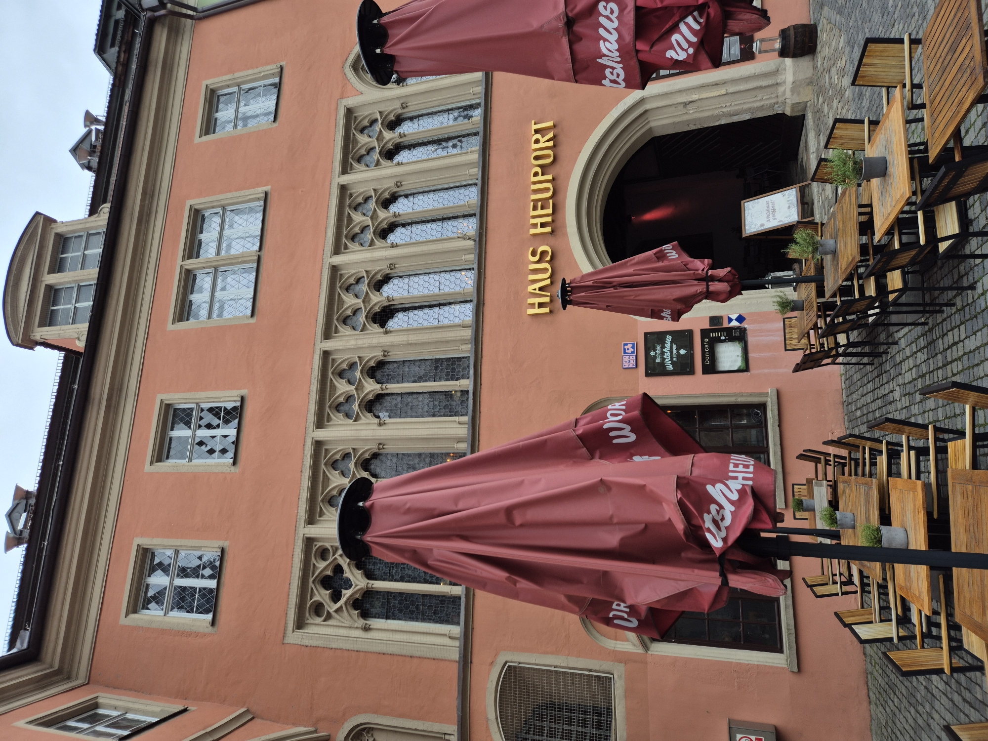 The image shows the exterior of "Haus Heuport" in Regensburg, Germany, a historic building with a distinctive salmon-pink facade. The upper floors feature ornate Gothic-style windows. In front of the building, there is an outdoor seating area for a restaurant or cafe, with numerous wooden tables and chairs arranged on a cobblestone pavement. Several large, closed red umbrellas are set up to provide shade. A sign above the arched entrance reads "HAUS HEUPORT" in golden lettering. Small signs advertising "Bischofshof Wirtshaus" and "DoniCafe" are visible on the wall. The scene appears to be a typical outdoor dining setting in a European city, with no people present at the time the photograph was taken. The sky is overcast, suggesting potentially inclement weather.