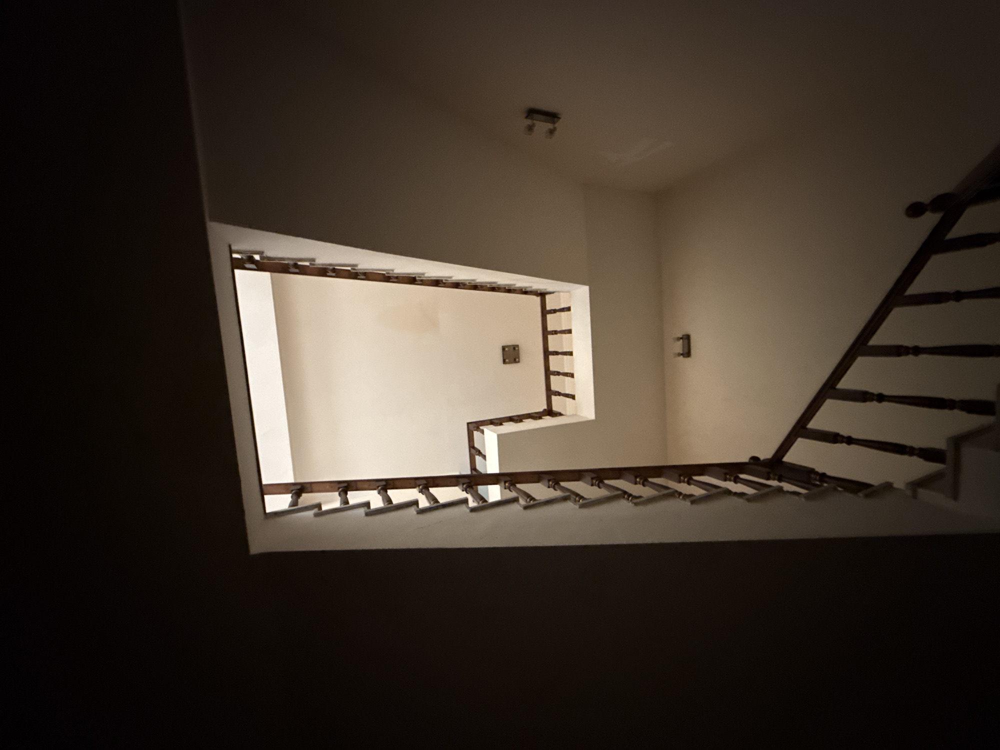 The image is an interior shot of a staircase in a building. The staircase has wooden banisters and appears to be multi-level, with the camera looking upwards. The walls are painted a neutral color, and there are light fixtures visible on the walls. The lighting is somewhat dim, casting shadows. The scene is devoid of any people or activity, presenting a static view of the architectural space. The location of Sabail Raion, Azerbaijan, is mentioned as context for this image.