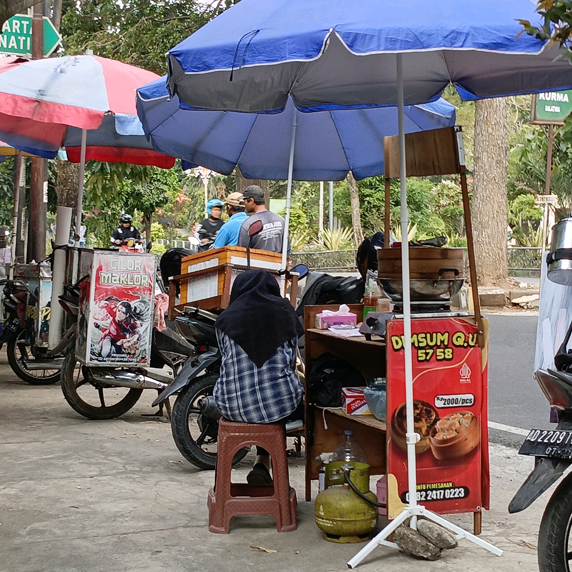 The image depicts a street scene in Salatiga, Indonesia, featuring a food stall. In the foreground, a person wearing a blue and white checkered shirt and a black headscarf is seated on a red plastic stool, facing away from the camera. To their right is a food cart with a red banner displaying "Dimsum Qu 5758" and an image of dim sum. The cart is equipped with a steaming apparatus. Behind the person, a motorcycle with a red and white graphic on its side, reading "CILOR MAKLOR," is parked. Several other motorcycles are visible in the background, along with other food stalls under blue and red umbrellas. Traffic signs indicating "ART. NATI" and "KURMA SALATIGA" are present. The environment suggests an outdoor market or roadside vendor area, with trees and greenery in the background. The ground is paved with concrete.