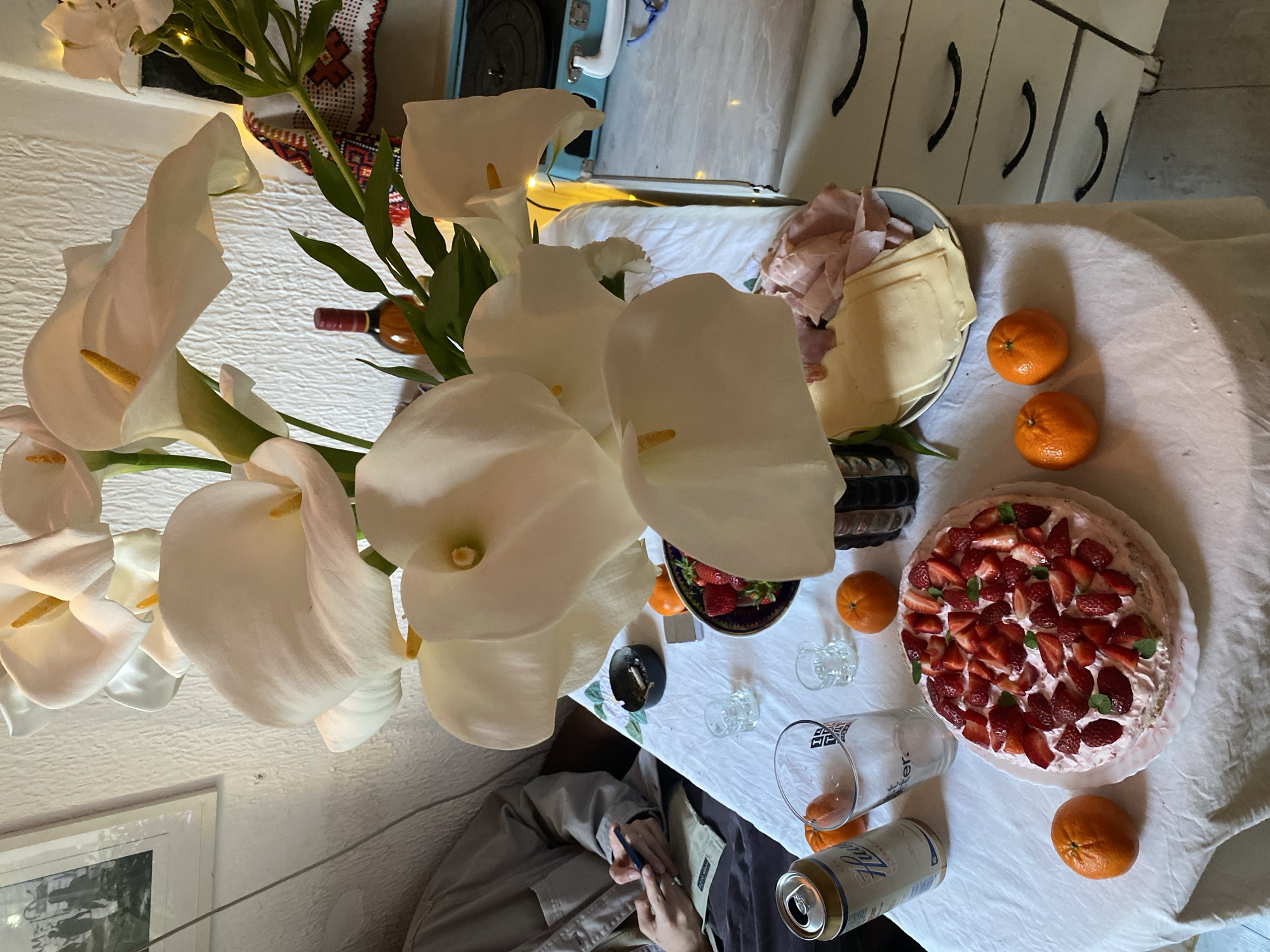 A high-angle shot captures a table laden with food and drinks, set against an indoor background. The table is covered with a white tablecloth. The scene includes a large bouquet of white calla lilies, a platter containing cheese and sliced ham, several oranges, a bowl of strawberries, a strawberry cake, beer in a glass, a can of beer, small glasses, and a small ashtray with a cigarette butt. A person, wearing a tan jacket, is seated at the table, holding a pen and notebook. In the background, elements of a room are visible, including white cabinets, a small blue appliance, and a framed picture on the wall. No specific details indicate that the location is in Belgrade, Serbia.