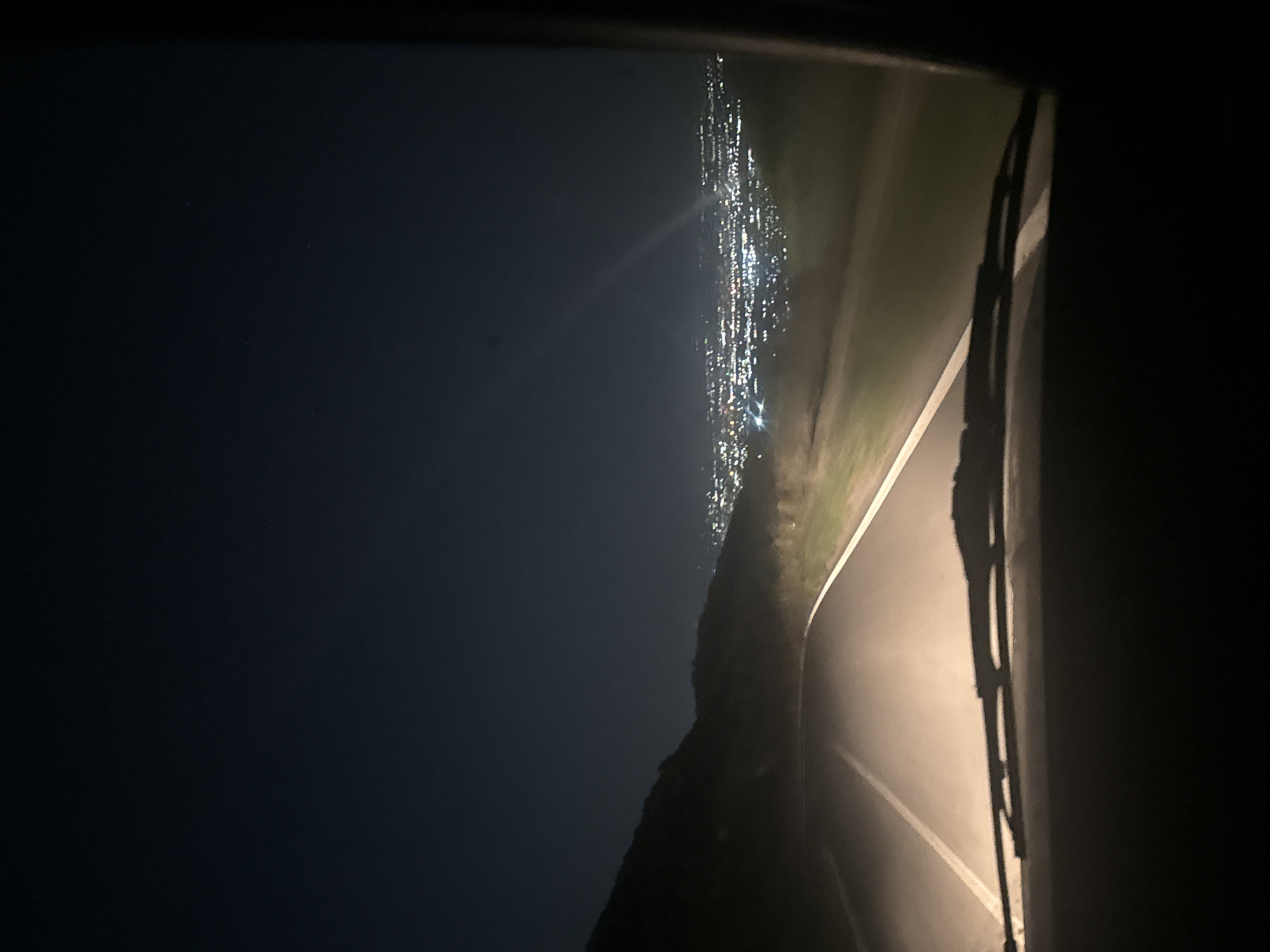 The image shows a view from inside a vehicle at night, looking out through the windshield. The vehicle is on a road that curves to the right. The headlights of the vehicle illuminate the road ahead. To the left of the road, a dark, silhouetted hill or mountain is visible. In the distance, a city is illuminated by countless lights, indicating a populated area. The sky above is dark blue, suggesting nighttime. The city lights stretch across the horizon, with some brighter points suggesting specific buildings or clusters of activity. A faint light streak or atmospheric effect is visible in the sky over the city. The foreground shows the lower edge of the windshield and the windshield wipers. The overall scene depicts driving through a rural or mountainous area towards a brightly lit city at night. The location is identified as Galabovo, Bulgaria.