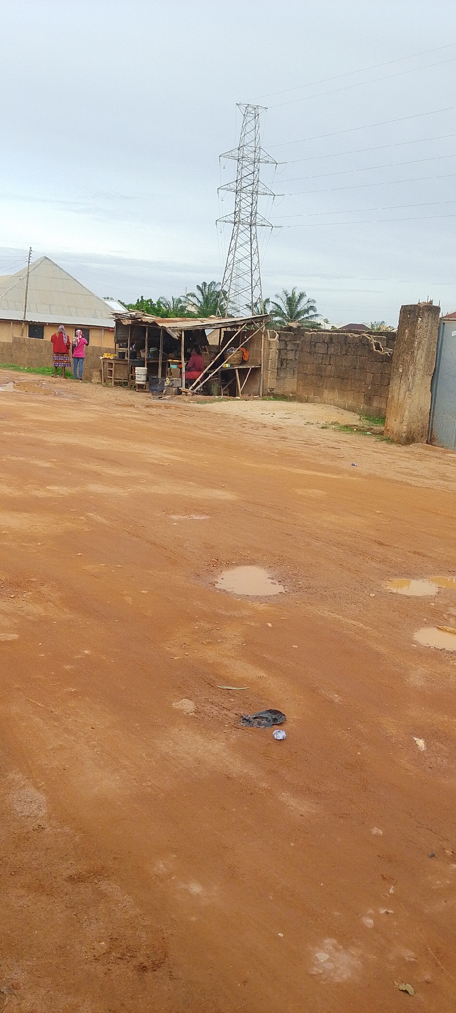 In the city of Jos, Nigeria, the image captures a street scene with a dirt road in the foreground. The road is uneven and contains several puddles of water. In the middle ground, a small roadside stall is visible with a few individuals present. One person, wearing a red top and a headscarf, is seated inside the stall. Two other individuals, one in a red top and another in a pink top and jeans, are standing outside the stall. The stall appears to be stocked with various items, and a large plastic container is placed near it. Behind the stall, a brick wall surrounds the area, and beyond that, palm trees and other vegetation are present. In the background, a tall electricity pylon dominates the skyline, with power lines extending from it. A light-colored, pyramid-shaped building with a corrugated roof is situated to the left of the stall, and another building with a similar roof is visible further in the distance. The sky is overcast, suggesting cloudy weather. A black plastic bag and a white object are on the ground in the foreground.
