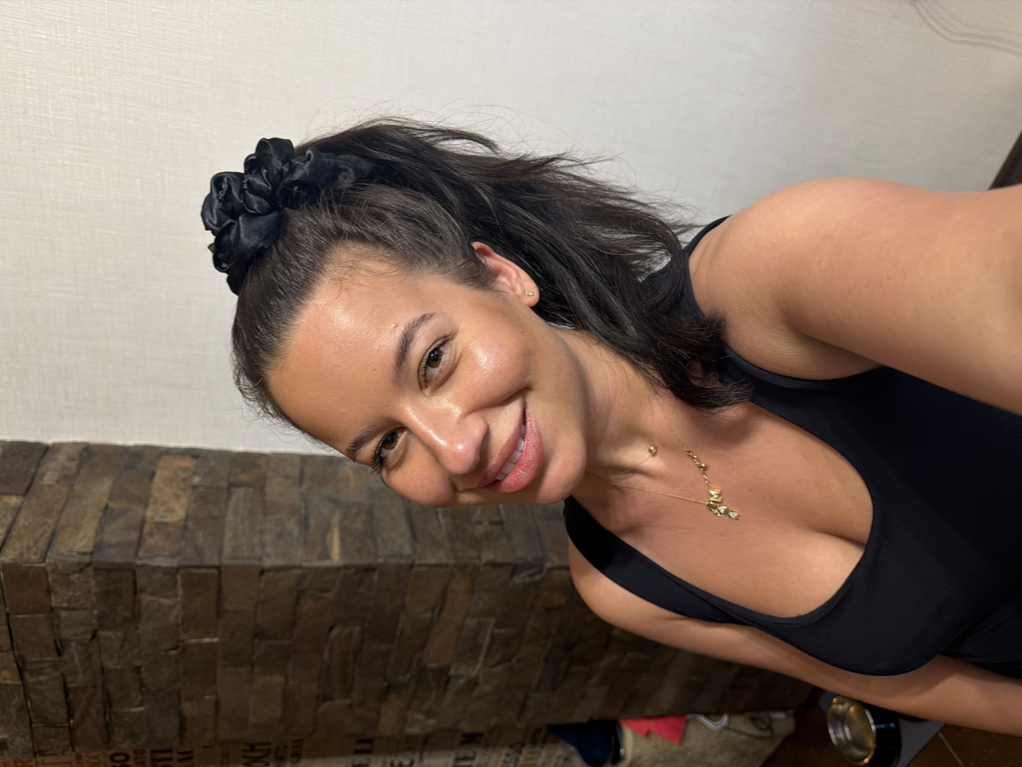 The image shows a woman with dark hair, styled in a high ponytail with a black scrunchie, and wearing a black tank top. She is wearing a gold necklace with small charms. She has on a small gold stud earring in her visible ear. She is smiling and looking towards the camera. The background includes a textured stone wall and a plain white wall. There are also some objects visible in the background on the left, including what appears to be a metal bowl on the floor and some stacked items. The image does not contain any information that would allow for the determination of its location in Burgas, Bulgaria.