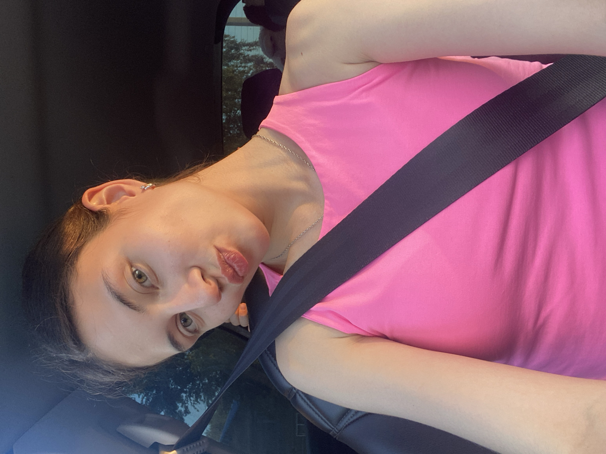 A young woman with brown hair and green eyes is in the driver's seat of a car, looking at the camera and puckering her lips. She is wearing a bright pink tank top and a silver necklace. A black seatbelt is across her chest. The sun is setting, casting a warm glow on her face. The background shows trees and a house, suggesting a suburban or rural setting. The car interior is dark. This image was taken in Votkinsk, Russia.