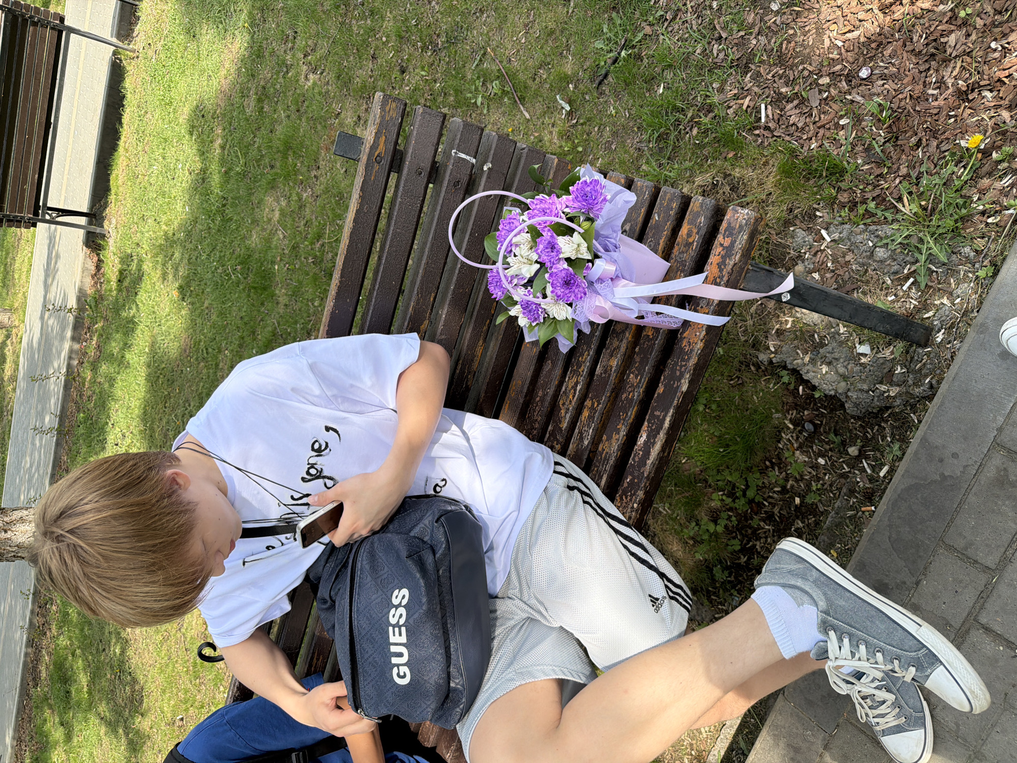 A high-angle shot depicts a person sitting on a wooden park bench. The person appears to be a young male with light blonde hair, wearing a white t-shirt, grey Adidas shorts, grey socks, and grey sneakers. The person is looking at a smartphone in their hands. A black GUESS brand bag is slung across their body. To the right of the person is a bouquet of purple and white flowers wrapped in purple tissue and ribbons, placed on the bench. The bench is located in a park with green grass and trees in the background. Part of another bench is visible on the left. The ground next to the bench is paved with grey paving stones. This appears to be in Chaikovsky, Russia.