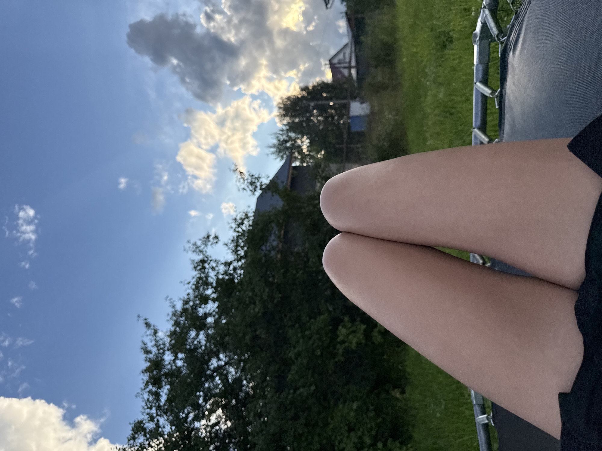 The image shows a close-up view of a person's legs, resting on what appears to be a trampoline. The person is wearing black clothing. The background consists of a rural landscape with green grass, trees, and houses, under a partly cloudy blue sky. The location is identified as Ветлужский, Russia.
