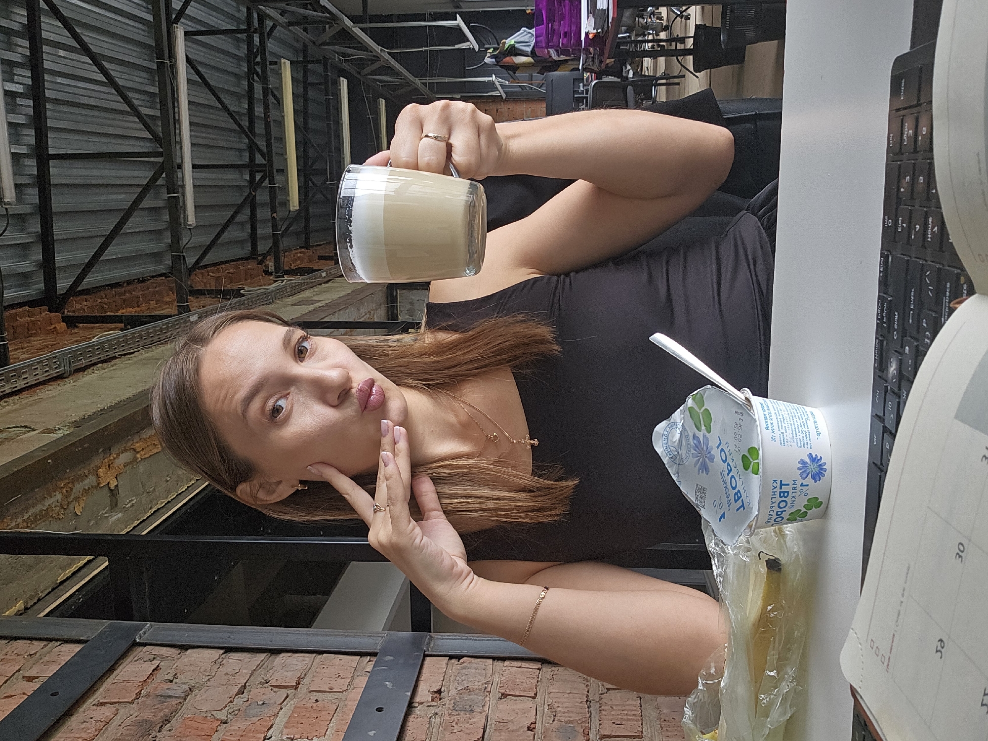 A young woman with long brown hair, wearing a black top, poses playfully in an industrial-style interior, likely a cafe or office in Chaikovsky, Russia. She is looking towards the camera with a slight pout, gently touching her cheek with her left hand. She wears a ring on each hand, a thin necklace with a small cross pendant, and a delicate bracelet.
With her right hand, she holds a clear glass mug containing a layered latte with distinct foam and coffee layers. On the white table next to her, an open white yogurt cup with a blue and green label is visible. Text on the yogurt includes "JBOL", "WOLKNN", and "KAHLABCKNN" (likely in Cyrillic). A banana wrapped in clear plastic sits beneath the yogurt. Part of a black keyboard and a notebook showing "30" and "es" are also on the table. The background features metal beams, corrugated metal walls, and brickwork, indicating a modern industrial design. The woman appears relaxed, enjoying a casual moment, possibly a coffee break.