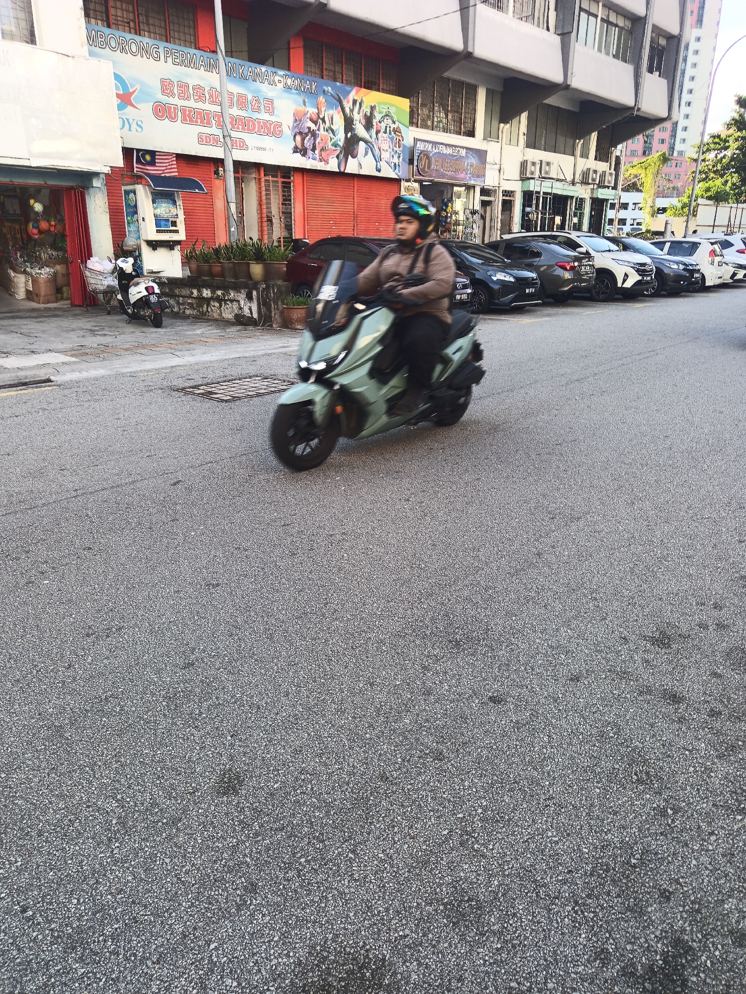 A person rides a light green scooter down a street in Kuala Lumpur, Malaysia. The person, wearing a helmet and a brown jacket, is in motion, as indicated by the blur of the scooter and the surroundings. The street is paved with asphalt and shows some wear. On the left side of the street, there is a building with a red facade featuring large signage in Malay and Chinese characters that appears to be a toy store. A white scooter is parked in front of the building. Several cars are parked on the right side of the street. The overall scene suggests a commercial area with moderate traffic.
