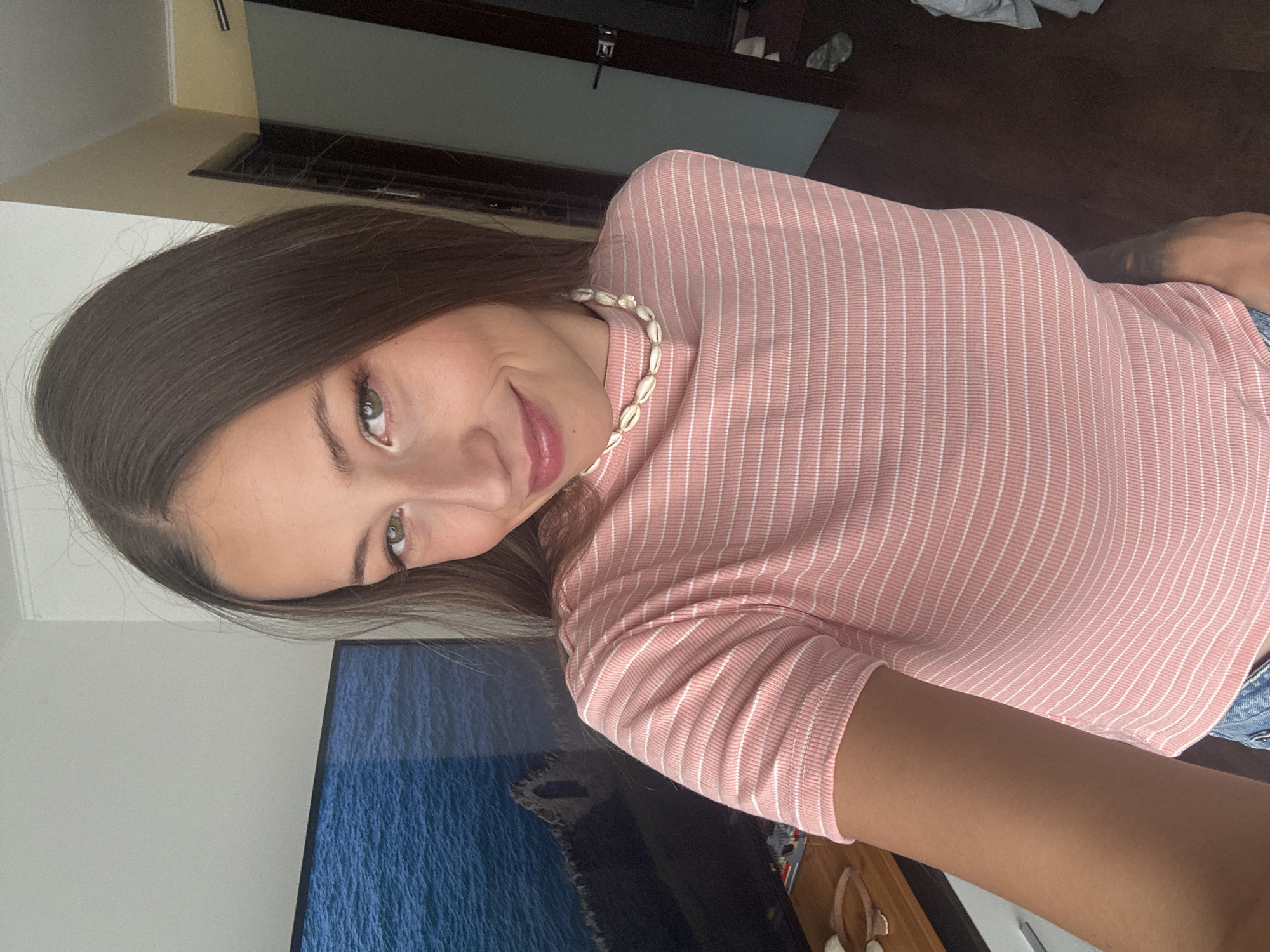 A woman with long brown hair and green eyes is posing for a selfie. She is wearing a pink and white striped shirt and a shell necklace. In the background, there is a television displaying a seascape, and a partially visible wooden door and some items are on a shelf. There is no indication of the location being in Saint Petersburg, Russia.