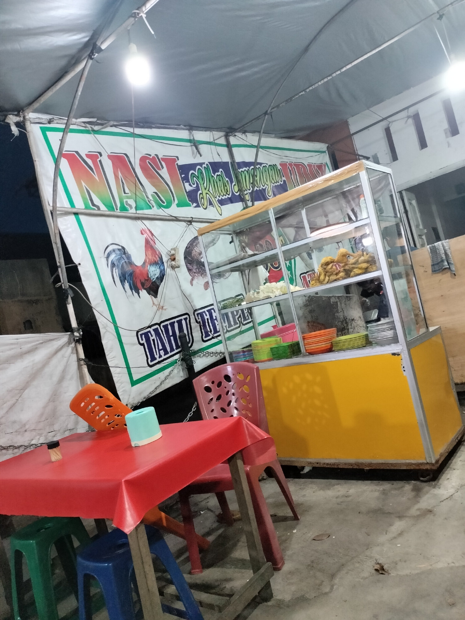 The image shows an outdoor food stall in Langsa, Indonesia. The stall features a large banner with colorful text that reads "NASI Khus Mengengan" and "TAHU TEMPE". A glass display case filled with fried food items and stacked plates is prominent. In front of the stall, a red plastic tablecloth covers a table with several plastic chairs of various colors (green, blue, and orange). A toothpick dispenser and a light blue cup are visible on the table. The ground is paved with concrete, and debris such as dried leaves are scattered on the surface. The stall is illuminated by a bright overhead light. The scene suggests a casual dining or takeaway food establishment operating in an urban outdoor setting.