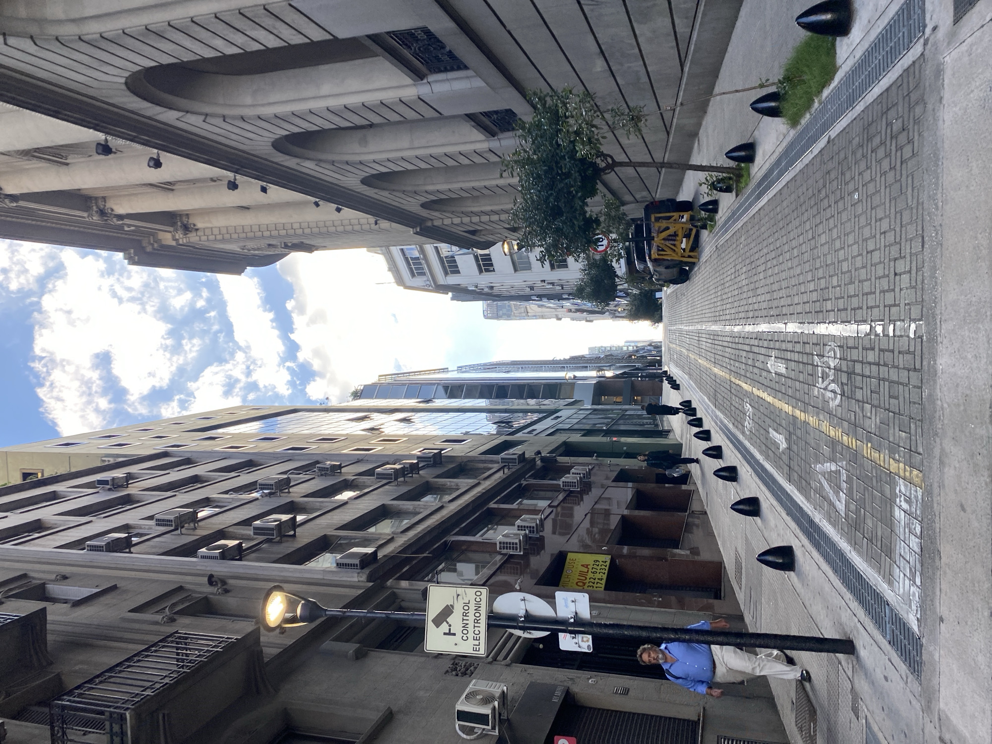 An upward-angled, tilted perspective of a narrow street lined by tall buildings in Buenos Aires, Argentina, under a sky partially covered by clouds. The street is paved with rectangular blocks and features a central yellow line marking the traffic direction. Black bollards are positioned along the edge of the sidewalk, separating the pedestrian zone from the road.
The buildings on either side showcase a variety of architectural styles. The building on the left has rows of windows and numerous air conditioning units, with a sign reading "CONTROL ELECTRONICO". At the base of this building, a man stands near a street lamp, wearing a blue shirt and khaki pants. The buildings on the right appear older, featuring arched windows and more ornate detailing.
In the distance, the street narrows further, and a yellow construction vehicle is visible. The trees are sparsely scattered along the street.