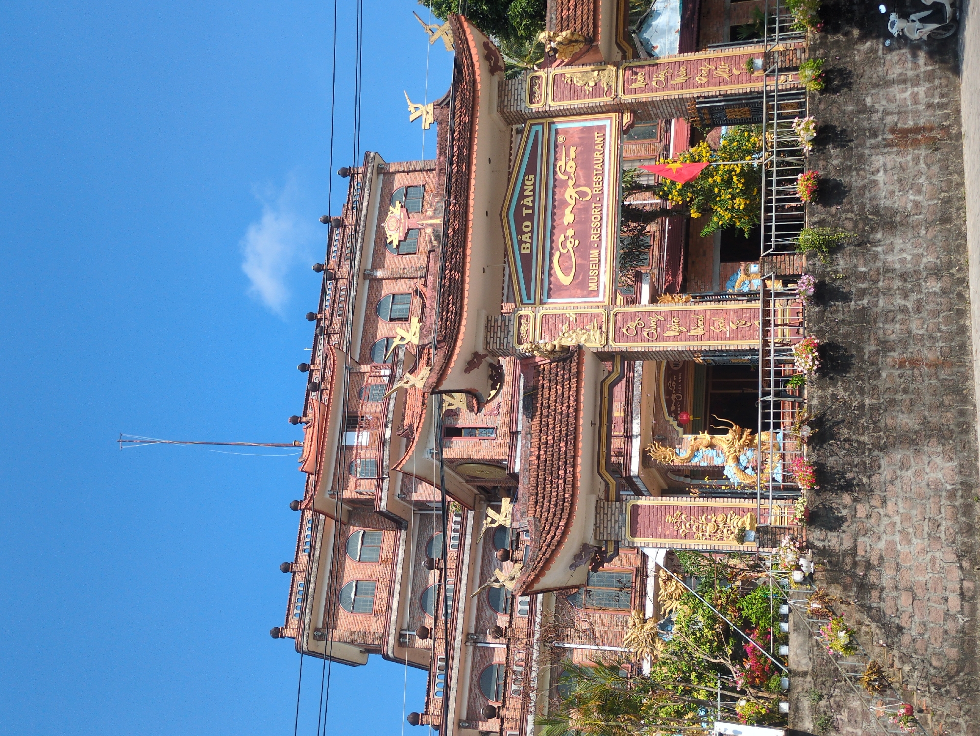 The image shows the ornate exterior of a building identified as "Bảo Tàng Cội Nguồn", which translates to "Roots Museum", a museum, resort and restaurant in Phú Quốc, Vietnam. The building is constructed of brick and has multiple stories with a red-tiled roof and a pagoda-style design. The architecture incorporates intricate carvings and decorative elements in gold and red. A Vietnamese flag is visible, as are potted plants adorning the building's perimeter. The sky is blue with a few clouds. Power lines are visible in the foreground. The building is situated next to a brick wall and walkway. A scooter is visible in the background.