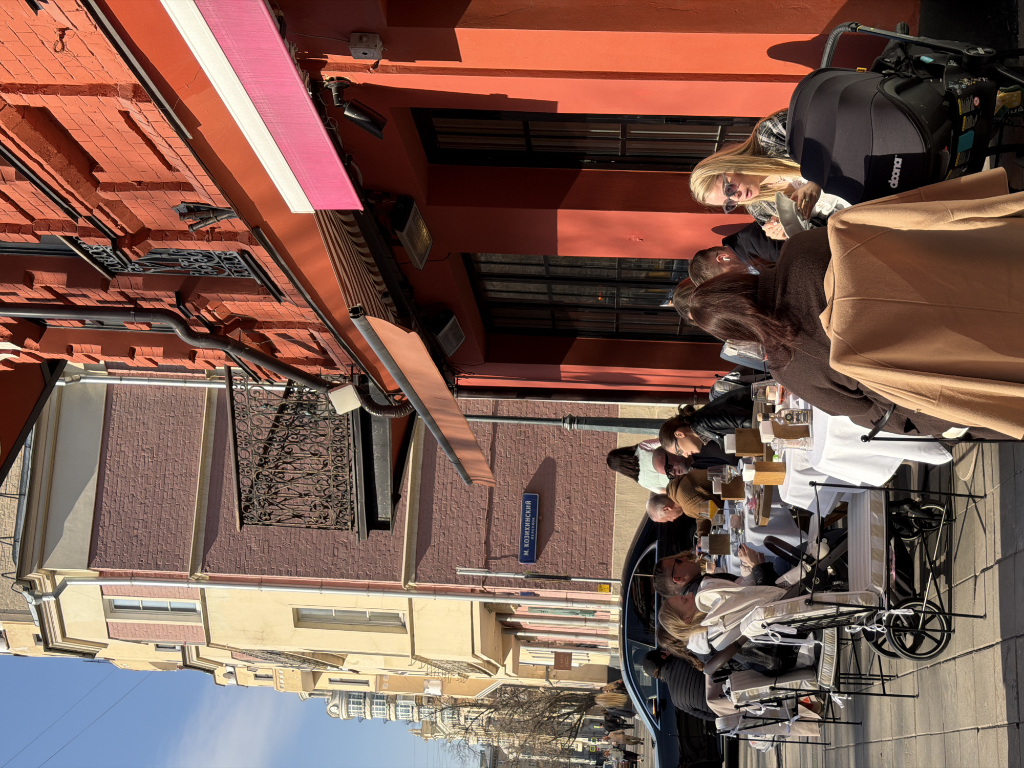 The image depicts an outdoor cafe scene on Maly Kozikhinsky Lane in Moscow, Russia, under a clear blue sky. The foreground features several tables with white tablecloths and dark-framed chairs with light cushions, arranged on a paved sidewalk directly in front of a building with a vibrant orange-red brick facade. Two awnings, one striped orange and white, and another pink and white, extend over the seating area, with outdoor heaters mounted to the building wall.
Approximately 10-12 individuals are seated at the cafe tables. At the rightmost table, a blonde-haired woman wearing sunglasses and a plaid shirt is looking into a white bowl, holding a utensil, positioned next to a black Doona brand baby stroller. Opposite her, a woman with dark hair is seen from the back, wearing a dark brown top, with a light brown coat draped over her chair. At an adjacent table, several people are engaged in conversation, including a blonde-haired woman in a white top, a man with dark hair and sunglasses, a bald man with sunglasses in a brown sweater, and a woman with dark hair and glasses in a black leather jacket. Various drinks, including what appears to be beer and orange juice, along with food items, are on their tables, accompanied by salt/pepper shakers and a bottle of spirits.
In the mid-ground, a dark-colored sedan is parked parallel to the street behind the cafe patrons. Pedestrians are visible walking on the sidewalk further down the street. The background features a multi-story building with a pale yellow and pinkish-red brick facade, featuring windows and a decorative black wrought-iron balcony. A blue street sign on this building clearly indicates "М. КОЗИХИНСКИЙ ПЕРЕУЛОК" (M. KOZIKHINSKY LANE). Strong sunlight creates distinct shadows across the scene.
