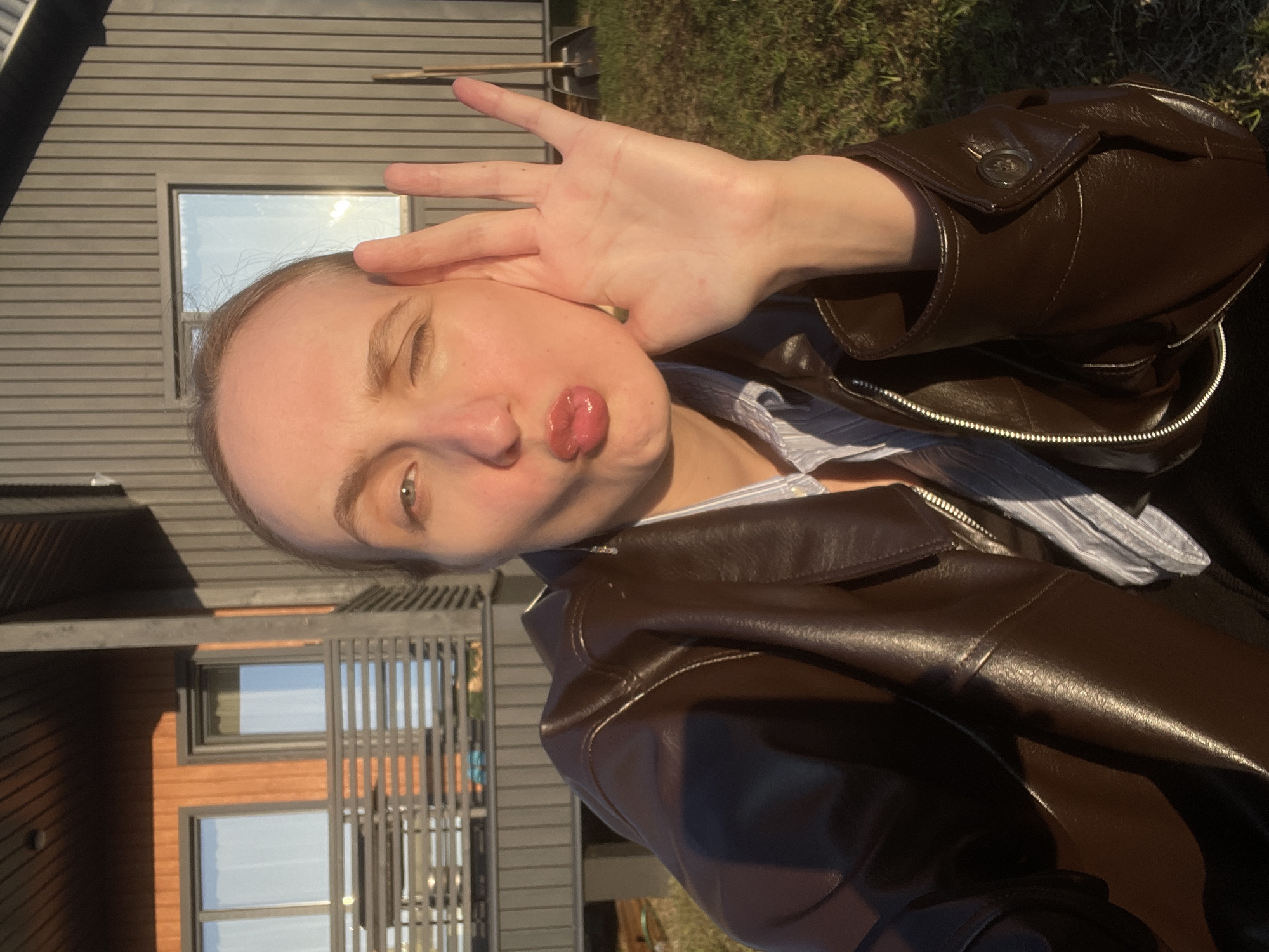 A selfie-style portrait, originally rotated 90 degrees clockwise, features a young adult positioned outdoors. The subject, appearing to be female with light skin and light brown hair, is captured from the chest up, making a playful "fish face" with puckered lips. Her right hand is raised with fingers spread, covering part of her face, possibly as a gesture or to block the sun. She wears a dark brown leather-look jacket over a light blue and white striped collared shirt.
The background showcases a modern building characterized by dark grey horizontal siding, interspersed with sections of light brown wooden paneling. Several rectangular windows in the building reflect a bright, clear sky. To the subject's right, a grey, slatted fence or pergola-like structure is visible. Green grass and foliage can be seen in the distant background.
The scene is bathed in bright, warm sunlight, indicating either morning or late afternoon, with highlights visible on the subject's skin and jacket. The overall atmosphere is casual and playful. No text is visible in the image. The setting suggests a contemporary residential or semi-rural environment, fitting with the location context of Sovetsko-Nikol'skoe Lesnichestvo, Russia.