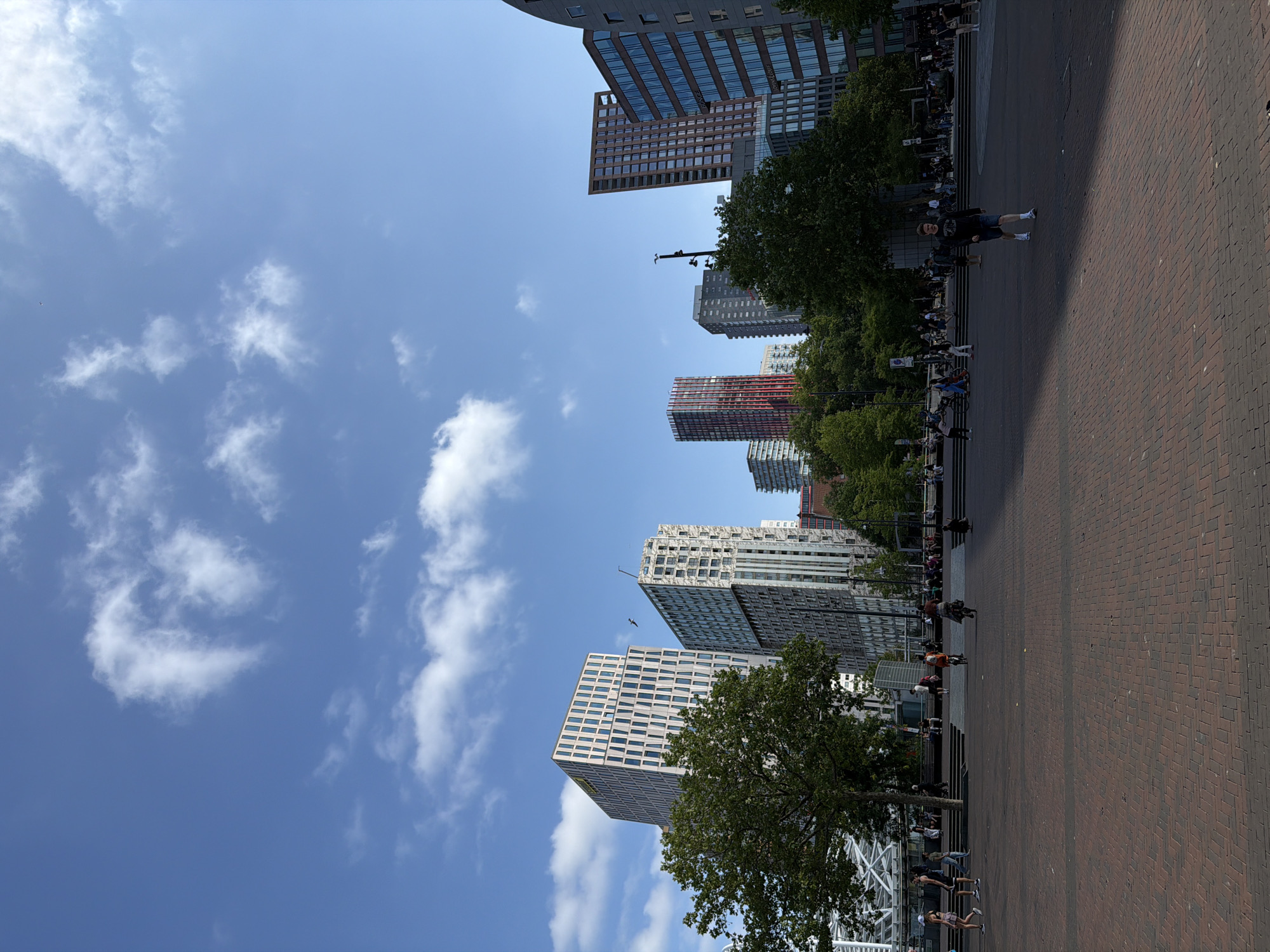 This is a street-level, medium shot of a large, open, paved square in front of several modern high-rise buildings in Rotterdam, Netherlands. The square is paved with reddish-brown brick pavers arranged in a herringbone pattern. The sky is a clear blue with scattered white clouds.
In the foreground, a young man wearing a dark sweatshirt, dark shorts, white socks, and white athletic shoes walks towards the camera from the right. He is walking in a patch of sunlight. In the middle ground, several people are walking, cycling, and standing around. Some are on bicycles, one of which is being ridden by a person wearing a helmet and a dark top. Further back, a group of people are seated on steps or benches in front of the buildings. A large green tree is visible on the left side of the image, partially obscuring two tall, modern buildings. One building has a white facade with a regular pattern of windows and vertical accents, while the other, to its right, has a grey, textured facade with a grid of windows.
In the background, a cluster of modern high-rise buildings dominates the skyline. These include a tall, slender building with a red facade and numerous windows, a reddish-brown rectangular building with many windows, and a large, modern building with a curved, grey facade and blue-tinted windows on the right. Trees are visible between the buildings and in the mid-ground, suggesting an urban park or green spaces integrated into the cityscape. Two birds are visible in flight against the sky, one near the building with the white facade and the other near the top of the building with the grey facade. The overall impression is a sunny, active urban environment.