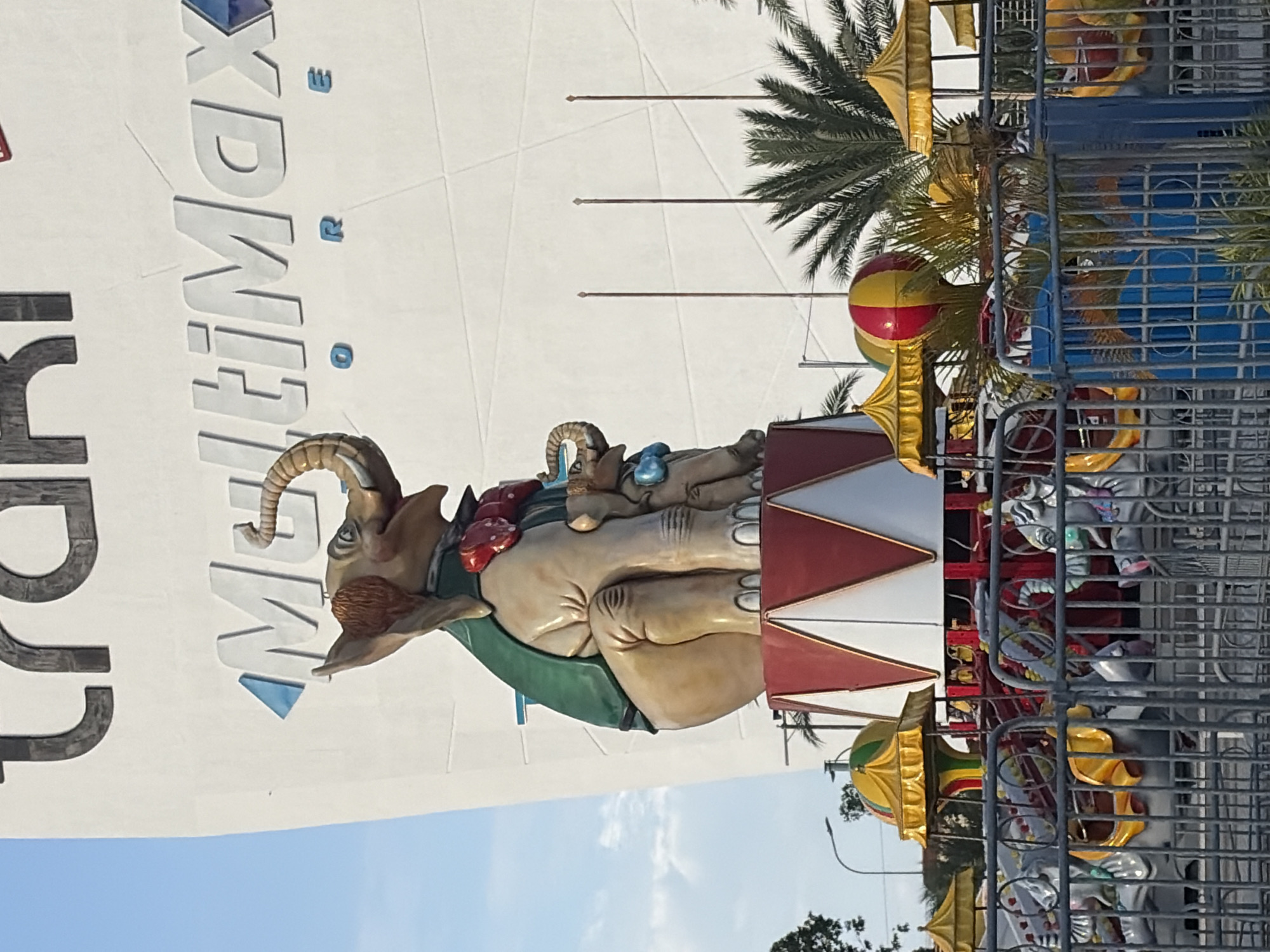 The image captures a carousel featuring an elephant sculpture, located at the MultiMax store in Ciudad Guayana, Venezuela. The carousel includes various other decorative elements like circus tents and other animal figures, all enclosed within a metal fence. Palm trees are visible in the background. The MultiMax logo is visible on a wall nearby. The sky is blue with light cloud cover.