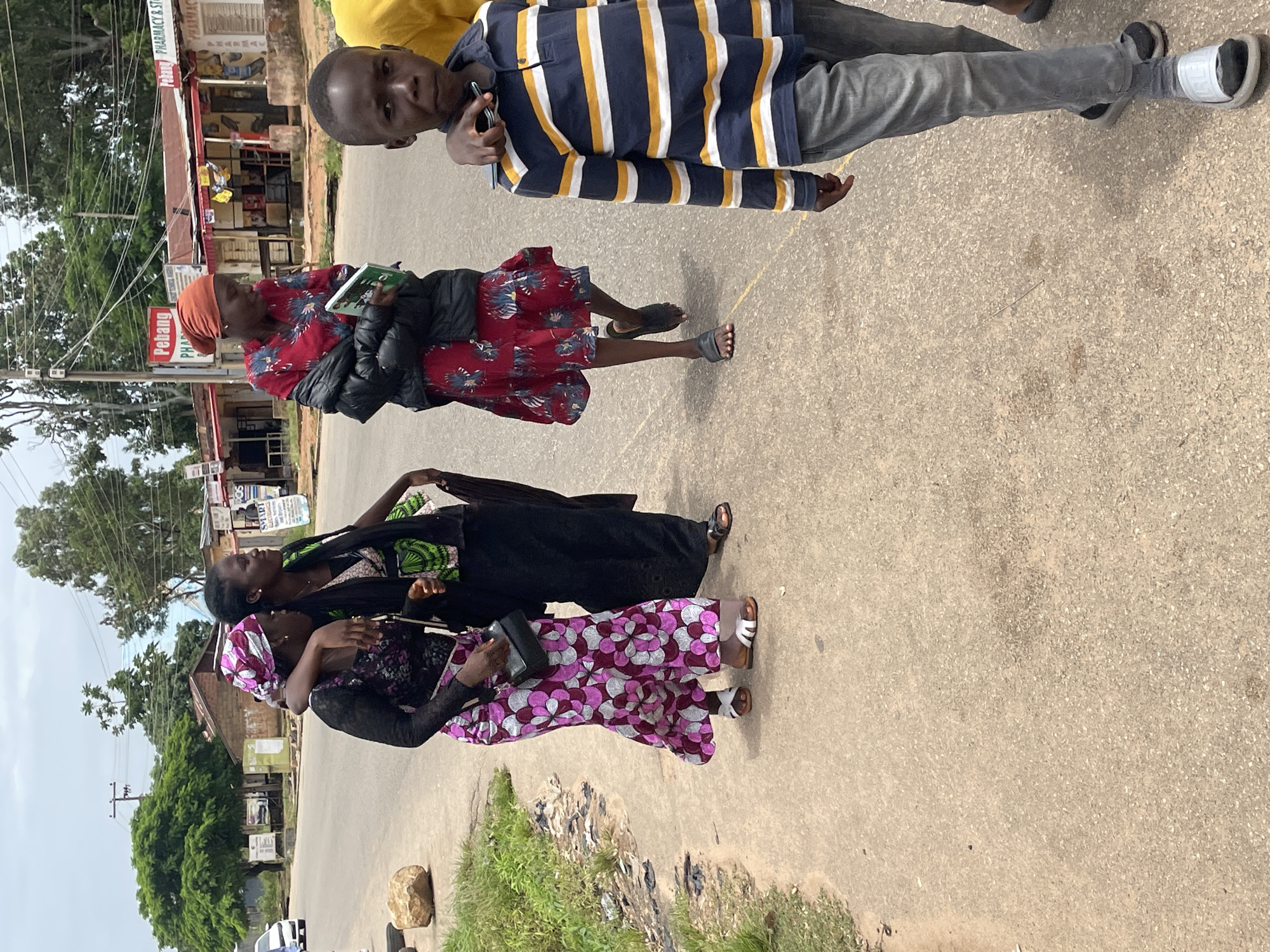 The image depicts a street scene in Jos, Nigeria, with a paved road and sidewalk. In the foreground, a young boy walks towards the camera wearing a striped shirt and grey pants. Behind him, three women are walking away from the camera. The woman on the far left wears a purple and white patterned dress and a headscarf. The woman in the middle wears a black dress with a green and black patterned overlay and holds a dark fabric. The woman on the far right wears a red patterned dress with a black jacket draped over her arm and holds a green book. The background shows buildings with shop fronts, trees, and a utility pole. The sky is overcast.
