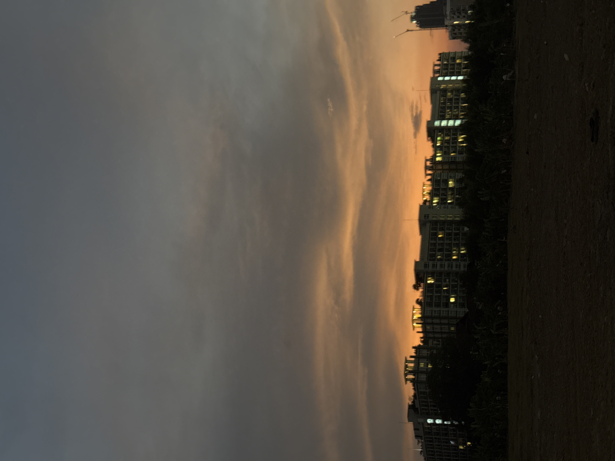 The image captures an urban landscape at dusk or dawn, showcasing a dramatic sky over a line of buildings. The sky, which dominates the upper portion of the frame, displays a gradient of colors, shifting from a deep, muted greyish-blue on the left to a warm, luminous orange and yellow on the right, indicative of a low sun angle. Thin, elongated clouds stretch across the sky, catching the soft light.
Along the horizon, a series of multi-story buildings, likely apartment blocks or commercial structures, are silhouetted against the vibrant sky. Many windows within these buildings are illuminated with bright, warm lights, suggesting an active urban environment as evening either begins or ends. Towards the far right, two prominent construction cranes are visible above the building line, signaling ongoing development, fitting for a growing city like Pattaya.
The lower part of the image features a dark, unlit foreground, appearing to be an expanse of unpaved ground or a grassy field. There are no discernible people or specific activities taking place, and no visible text. The scene conveys a tranquil yet dynamic urban setting during twilight, with the city's artificial lights beginning to contend with the fading natural light of the sky.