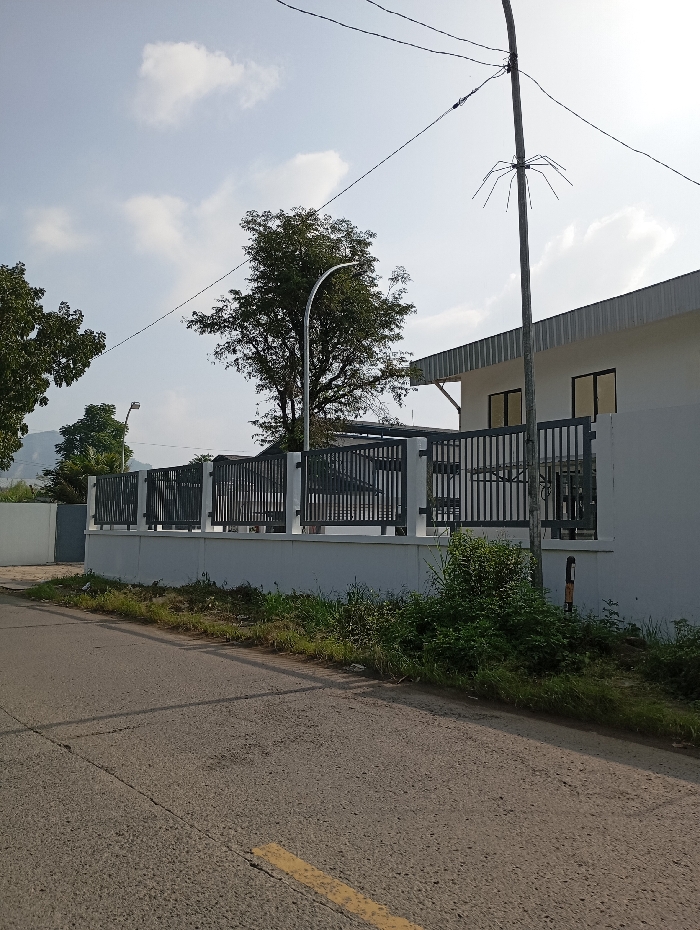 An exterior scene depicts a modern, single-story building situated along a paved road in Pataruman, Indonesia. The building features a light-colored facade with dark-framed rectangular windows and a silver-colored corrugated metal roof section on its right side. A dark-colored metal fence, comprising vertical bars atop a white concrete wall, encloses the property. The foreground consists of an asphalt road exhibiting cracks, with a faded yellow line visible on its left side. Green vegetation, including grass and shrubs, lines the edge of the road, bordering the property wall. Several trees with dense green foliage are visible behind the fence and in the background. A prominent light-colored utility pole stands on the right, supporting multiple dark utility wires. Another utility pole with a curved streetlight is visible further back on the left. The sky is clear blue with scattered white clouds, indicating daylight conditions. No individuals are present, and no actions are discernible within the frame.