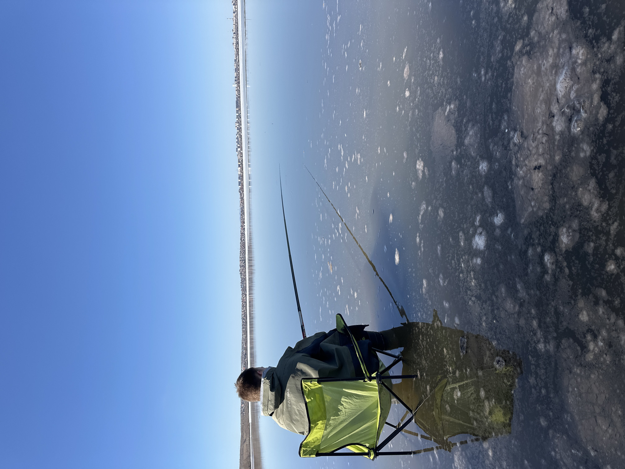 A person sits in a bright green folding chair on the edge of a body of water, possibly frozen, holding two fishing rods. A city skyline is visible on the far shore. The sky is clear and blue.