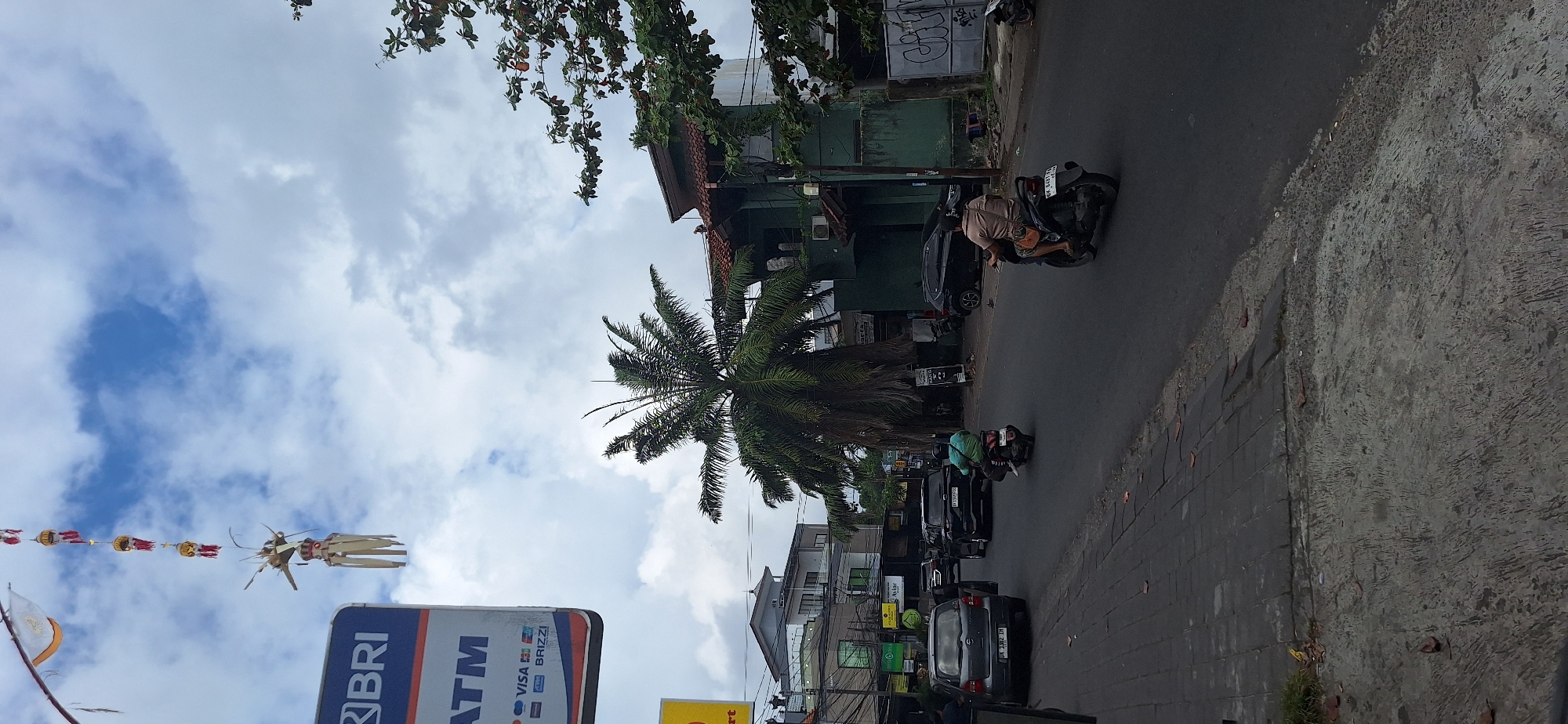 The image depicts a street scene in Kuta Selatan, Indonesia. A paved road occupies the foreground and extends into the mid-ground, with a gray, textured sidewalk bordering the left side. Several vehicles are present: a gray hatchback with license plate "DK 1382 IH" is parked on the left, followed by a black SUV, and another black car further down the road. Two individuals are riding motorcycles. One is a person in a blue and green shirt on a black motorcycle, moving away from the viewer on the road. Another individual on a brown motorcycle, wearing a light brown shirt, is also traveling away from the viewer.
In the background, a prominent tall palm tree stands out, its fronds reaching towards the sky. Behind the palm tree are several buildings, including a multi-story structure with a gray roof and balconies, and a darker green building with a tiled roof. A sign with "BRI" and "ATM" in blue and white is visible on the left side of the image.
Decorations hang from a pole on the left, featuring red and white ornaments and what appear to be woven palm leaf elements. The sky is filled with white and gray clouds. The overall environment suggests a commercial or residential street with moderate traffic.