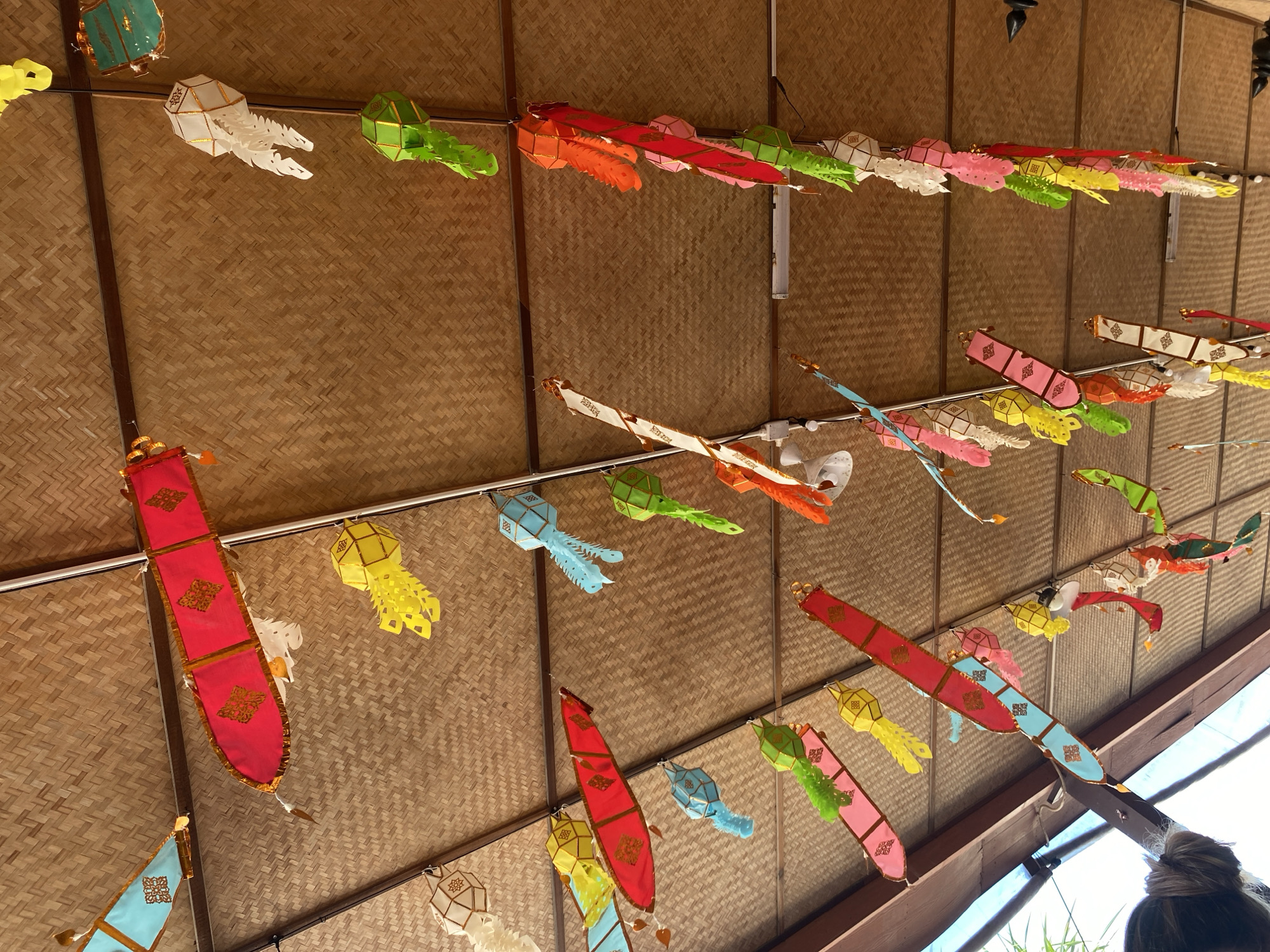 The image shows a ceiling decorated with numerous colorful paper lanterns. The lanterns are of various shapes and sizes, including cylindrical, hexagonal, and teardrop shapes, and are predominantly in shades of pink, red, yellow, green, blue, and white. Some lanterns have intricate patterns or cutouts. They are suspended from the bamboo-paneled ceiling by thin strings. The lighting suggests it is daytime, with natural light filtering in from an unseen source. The overall impression is festive and decorative. The scene is located in Baan Lamai, Thailand, likely a public space or market given the extensive decoration. No people are clearly visible except for the back of a person's head and shoulders in the lower left corner of the frame, appearing to look up at the decorations.