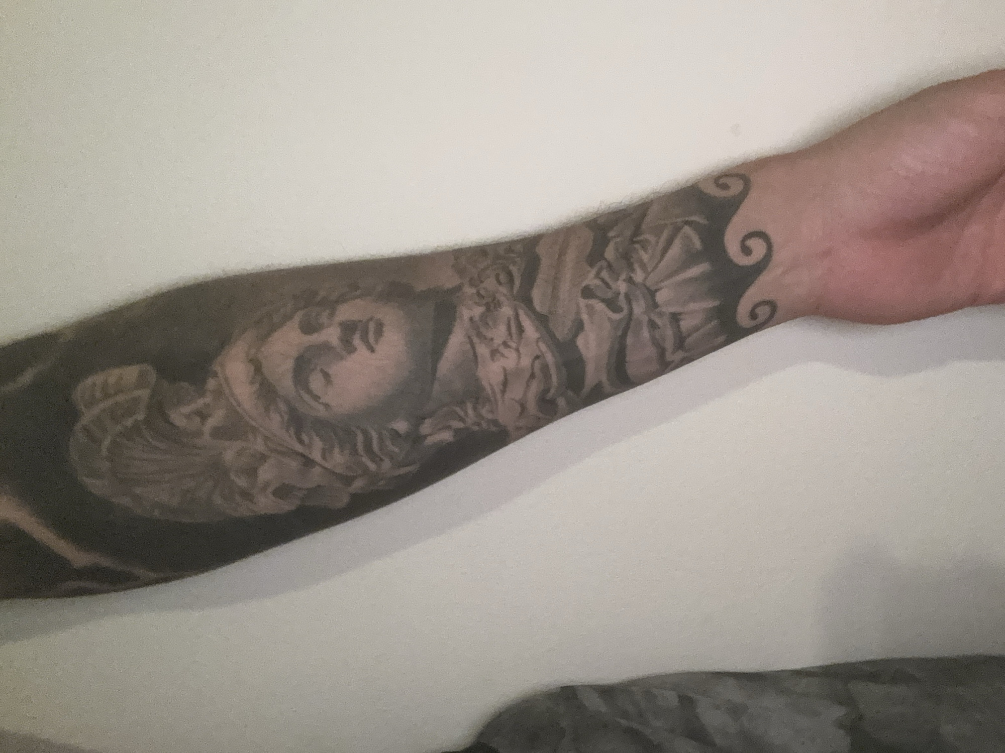 A grayscale tattoo of a woman's face and figures adorns a person's forearm. The tattoo extends from the wrist to the lower arm, with swirls at the wrist area. The background is a plain white wall. An indistinct dark object is visible at the bottom of the image. The scene is indoors. There is no indication of the city of London in the image.
