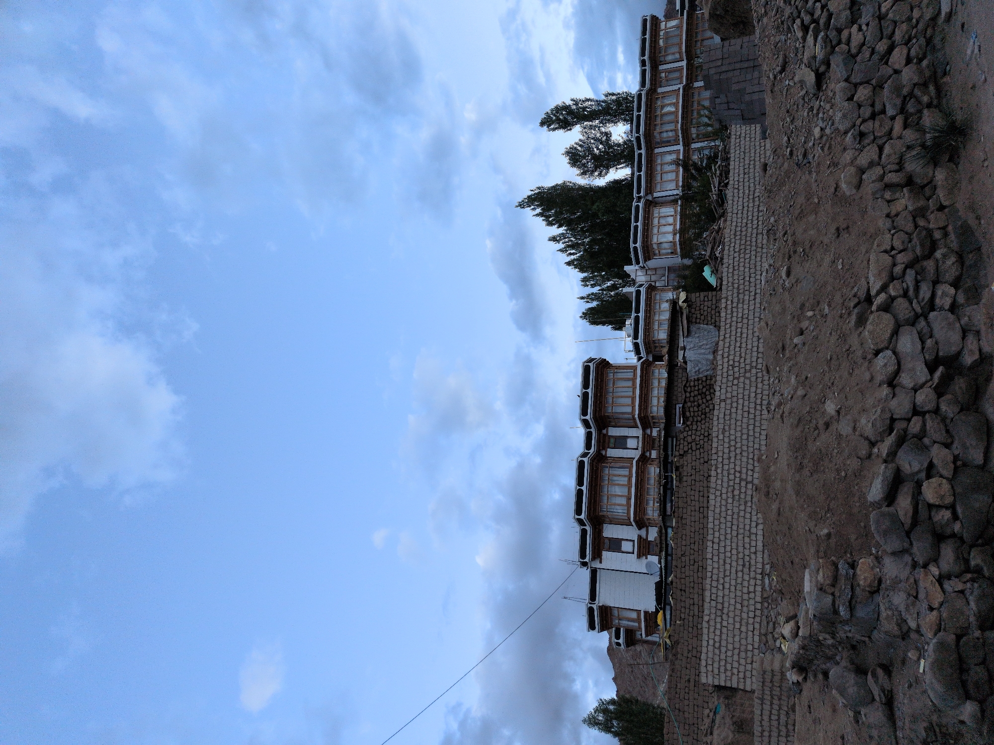 The image depicts a multi-story building with white walls and wooden-framed windows, situated on a hillside in Leh, India. The building features prominent bay windows and a dark-colored roofline. In front of the building, there is a brick wall and a sloped area covered in dirt and rocks. Several trees are visible behind and to the side of the building, with a mountain range faintly visible in the background. The sky is a pale blue with scattered clouds. A single, thin wire stretches diagonally across the upper left portion of the frame. The scene appears to be an outdoor, daytime shot, possibly taken during early morning or late afternoon given the lighting.
