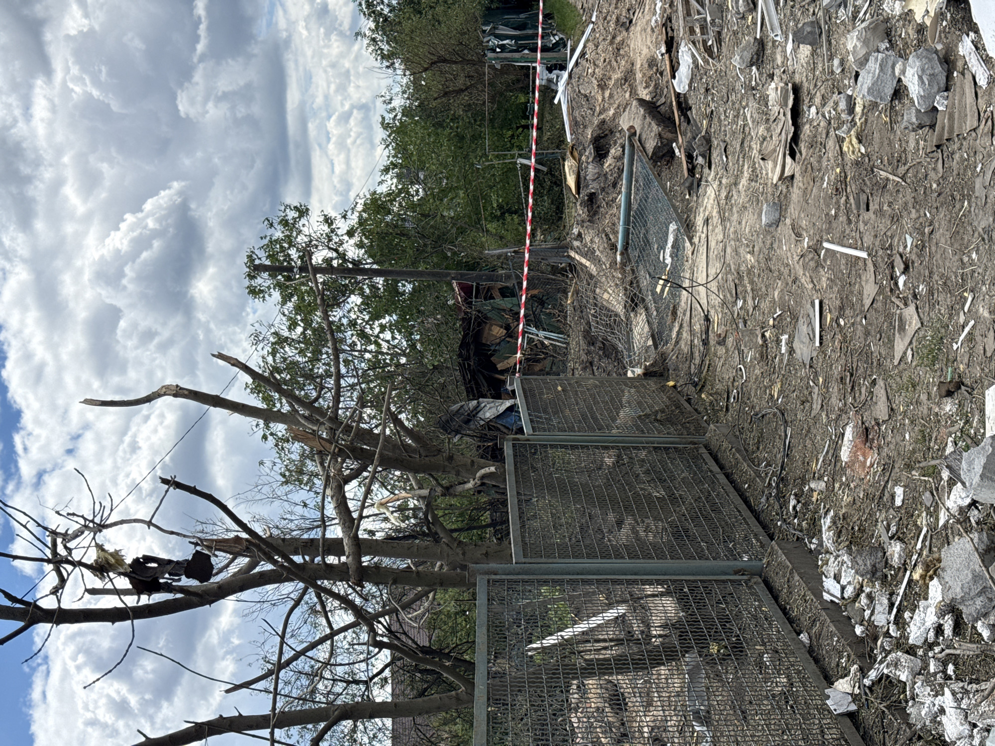This image depicts the aftermath of an apparent explosion or attack in Kyiv, Ukraine. A metal mesh fence, partially damaged and leaning, runs across the foreground. Beyond the fence, the scene is one of significant destruction. Trees are broken and stripped of foliage, with debris scattered around their bases. A red and white striped tape, commonly used for caution or cordoning off an area, is stretched across the mid-ground, further indicating a hazardous zone. The ground is covered in rubble, including pieces of concrete, metal sheeting, and various other fragments. In the background, the remains of structures, possibly sheds or outbuildings, are visible, some appearing collapsed or heavily damaged. Green foliage from surrounding trees and bushes provides a stark contrast to the destruction. The sky is partially overcast with large, white clouds. There are no people visible in the frame.