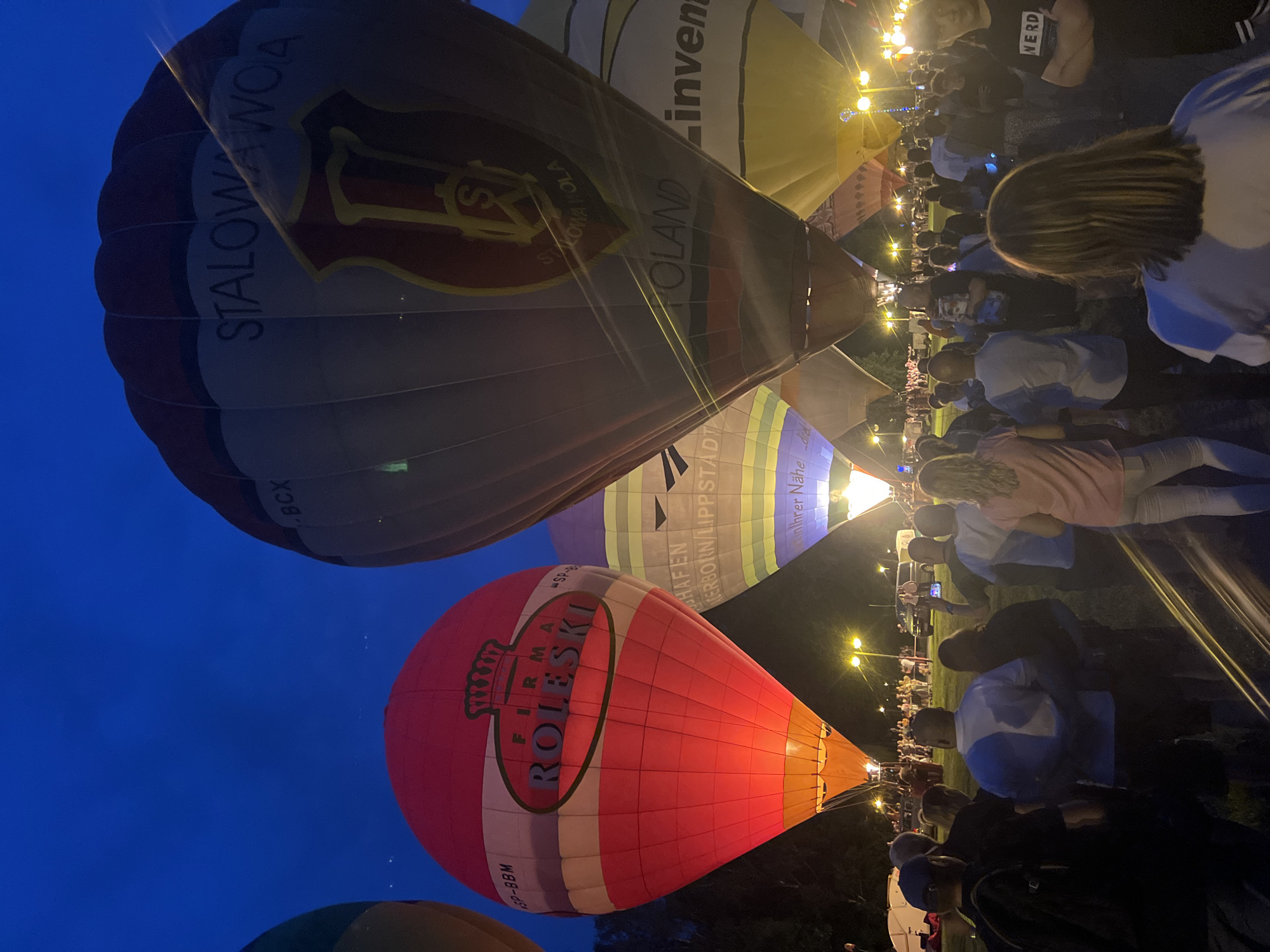 The image depicts a hot air balloon festival at dusk in Tychy, Poland. The foreground is filled with a crowd of people facing away from the camera, looking towards several illuminated hot air balloons. The balloons are in various colors and designs, with some displaying text and logos. One prominent red and white balloon in the center has "FIRMA ROLESKI" written on it, along with a crown emblem. Another large balloon to the right features the text "STALOWOWA WOLA" and a shield with an "S" logo. In the background, more balloons and lights are visible, suggesting a large event. The sky is a deep blue, indicating the transition from daylight to night. The environment appears to be an open field with some trees and lampposts visible. The people in the foreground are casually dressed, and their postures suggest they are observing the spectacle.