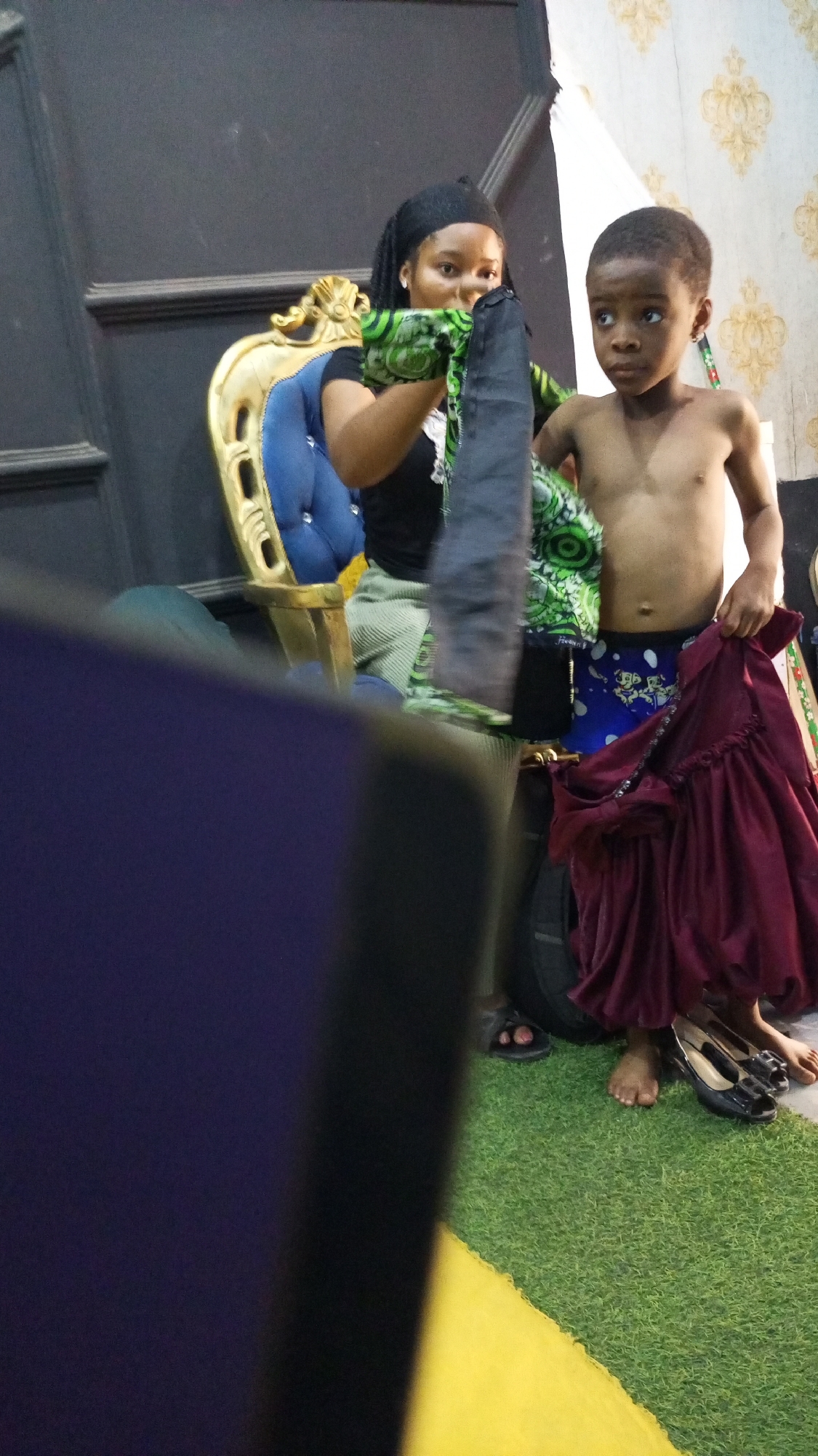 A woman and a child stand in front of a wall decorated with a patterned wallpaper, in what appears to be a photography studio with a black panel, ornate gold and blue chair, and artificial green grass. The woman, wearing a black top, striped pants, and a head covering, holds up a colorful fabric. The bare-chested child, wearing patterned shorts, holds a maroon dress. Women's shoes are visible on the artificial grass.