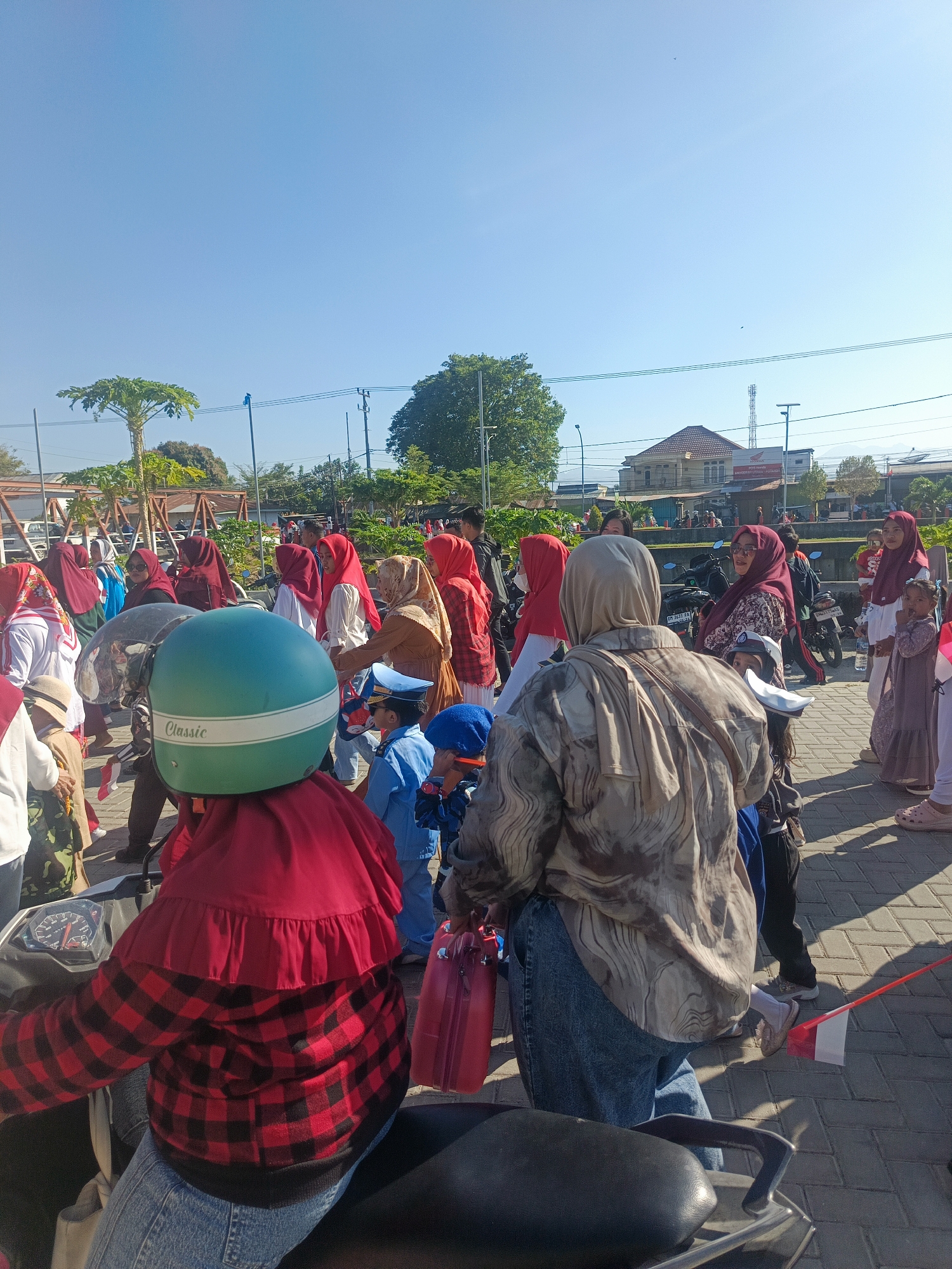 The image depicts a sunny outdoor scene with a group of people, many wearing hijabs, gathered on a paved walkway. In the foreground, a person is seated on a motorcycle, wearing a red and black plaid shirt and a red hijab. A prominent green helmet with a white stripe and the word "Classic" is placed on the motorcycle's seat. A child in a blue uniform and hat stands to the right of the motorcycle. Several Indonesian flags, red and white, are visible throughout the scene. In the background, buildings and trees are present, and power lines crisscross the sky. The overall atmosphere suggests a public gathering or event. The presence of Indonesian flags and the general setting indicate this location is likely in Indonesia, and given the user's prompt, it is specified as being in the city of Gorontalo, Indonesia.