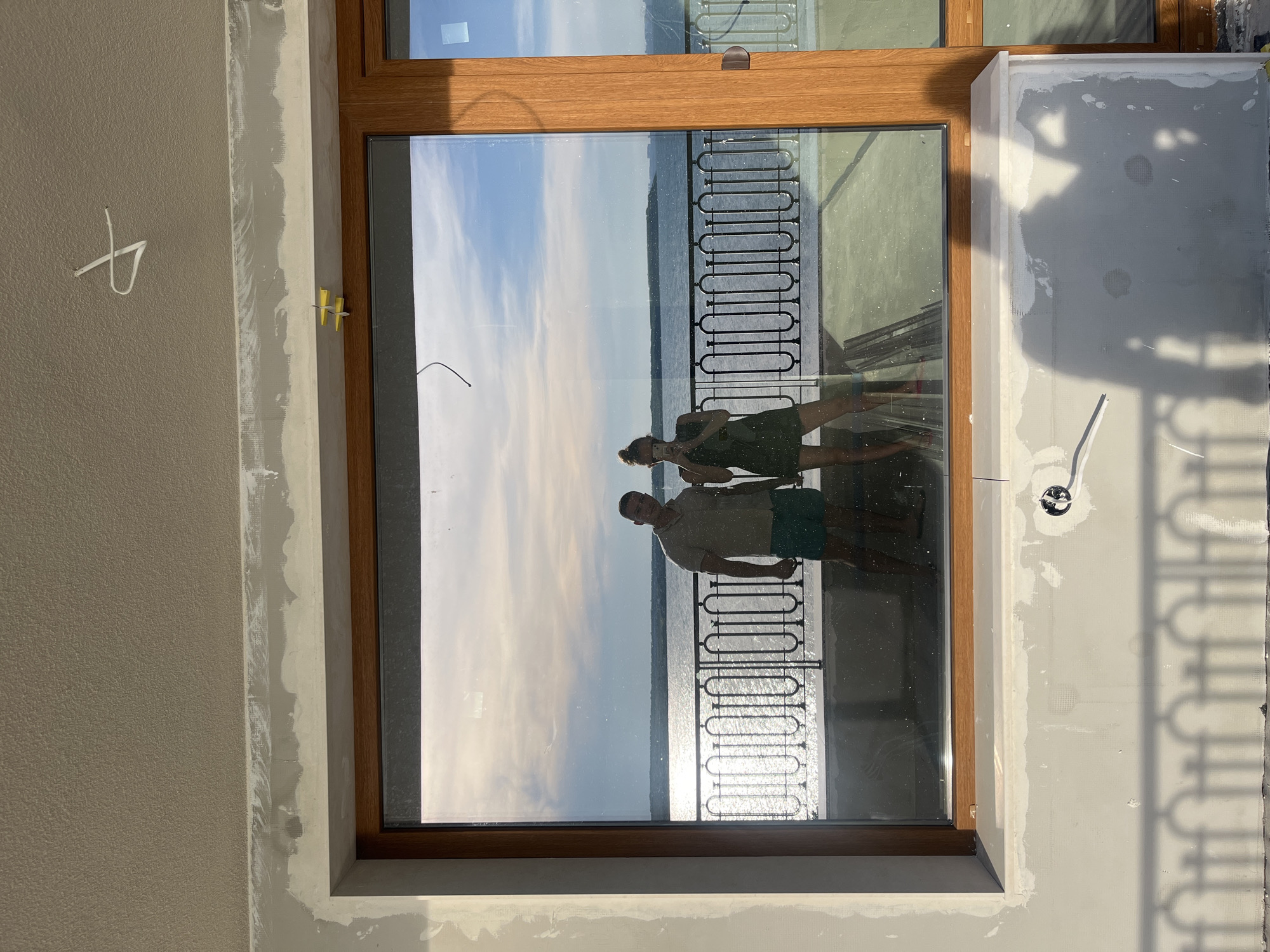 The image shows a reflection in a large window of two people standing on a balcony overlooking the sea. The man on the left is wearing a light-colored collared shirt and turquoise shorts. The woman on the right is wearing a dark green dress and has her hair in a bun. She is holding a phone and appears to be taking a photo of herself and the man. In the background, the sea stretches out to the horizon under a partly cloudy sky. The balcony has a black metal railing with an ornate pattern. The area around the window is under construction, with unfinished plaster on the walls and ceiling. There are yellow construction wedges visible above the window frame. A white wire hangs down from the ceiling in the upper right corner of the image. The scene is likely located in Pomorie, Bulgaria, given the architectural style of the railing and the presence of the Black Sea.