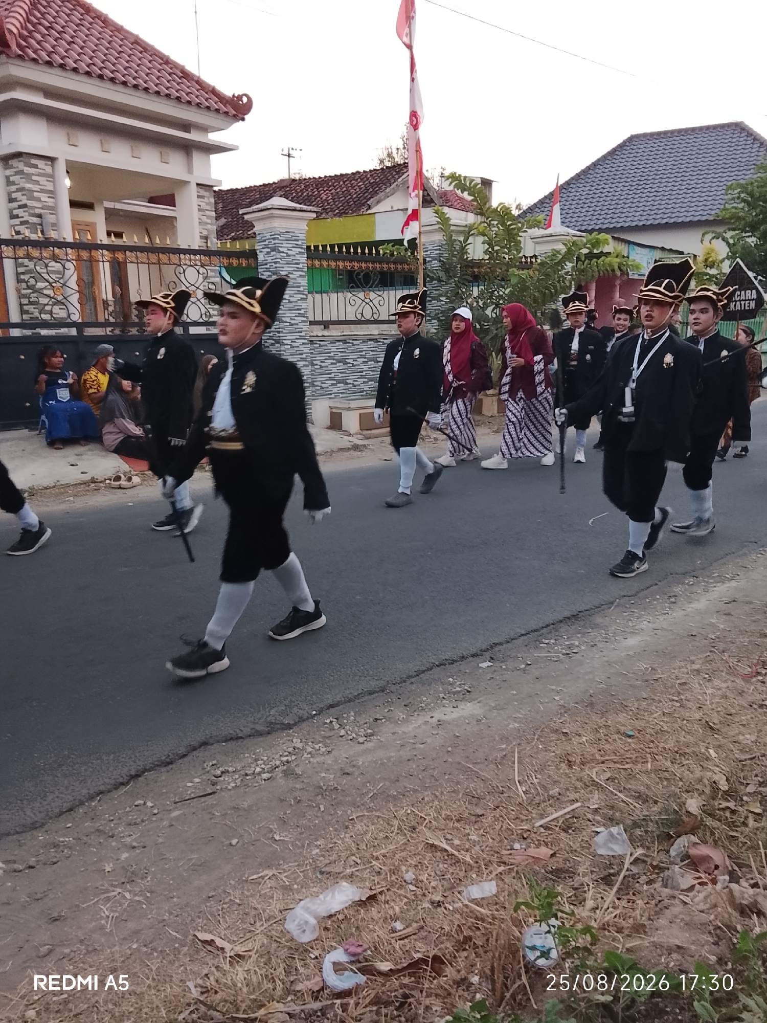 A group of young males in uniform, possibly a marching band or ceremonial group, walks down a paved road in Semanding, Indonesia. They are dressed in black jackets with white shirts and ties, black pants, white socks, and distinctive black hats with gold trim. Some carry batons or staffs. They are accompanied by a few women in traditional Indonesian attire and a flag bearer with a red and white Indonesian flag. The scene appears to be a procession or parade, with houses visible in the background. The environment is a residential area with some vegetation and scattered debris on the roadside.
