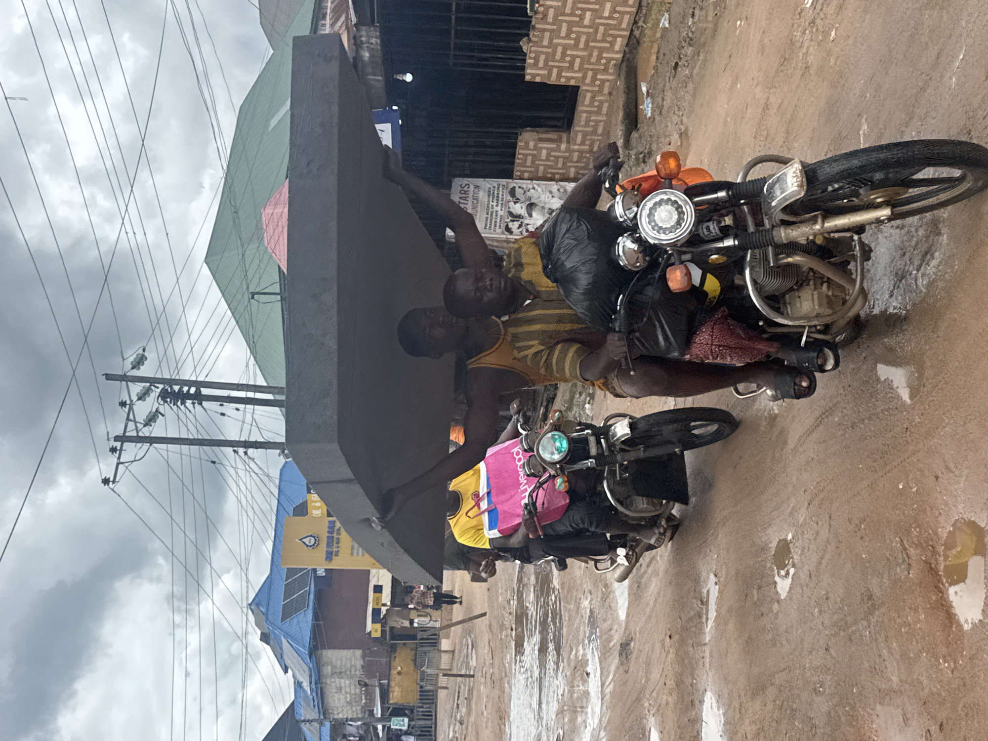 In Ihiagwa, Nigeria, two men transport a large, dark, flat object, possibly a slab, on the back of a motorcycle. The man in the front, wearing a yellow and striped shirt, drives the motorcycle. The man behind him, in a yellow tank top, balances the object on his head and shoulders. A third person, partially visible on the left, rides another motorcycle behind them, also appearing to carry a person. The scene unfolds on a muddy, wet road with several puddles, suggesting recent rainfall. Buildings line the street, some with corrugated metal roofs, and power lines crisscross overhead. A sign for "STARS COMMUNICATION" is visible on one of the buildings. The overall atmosphere is one of everyday life and resourcefulness in an urban environment.