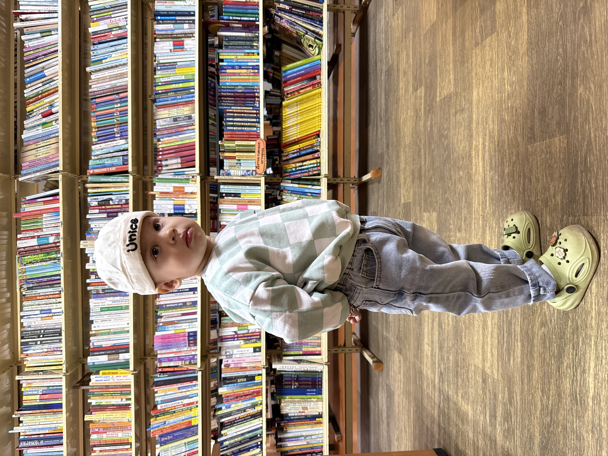 A young child is captured standing sideways in what appears to be a library or bookstore, looking directly at the camera with a curious expression. The child wears a light-colored flat cap with the word "Unice" visible on the front, a light green and white checkered long-sleeved shirt, light blue jeans, and light green clogs. One of the clogs features a charm partially reading "HAPPY HUGS" and another depicting a basketball.
The background is filled with tall, densely packed bookshelves, brimming with various books of different sizes and colors, stacked both vertically and horizontally. The shelves appear to be made of light-colored wood or metal. Visible text on one book spine, seen in a cropped image, reads "Детективные истории" (Detective Stories) in Cyrillic. The floor beneath the child is made of light to medium brown wooden planks. The setting is indoors, with ambient lighting, suggesting a cozy and book-filled environment, possibly in Rusanovo, Russia. The child's hands are lightly clasped in front, and their posture is upright and engaged.