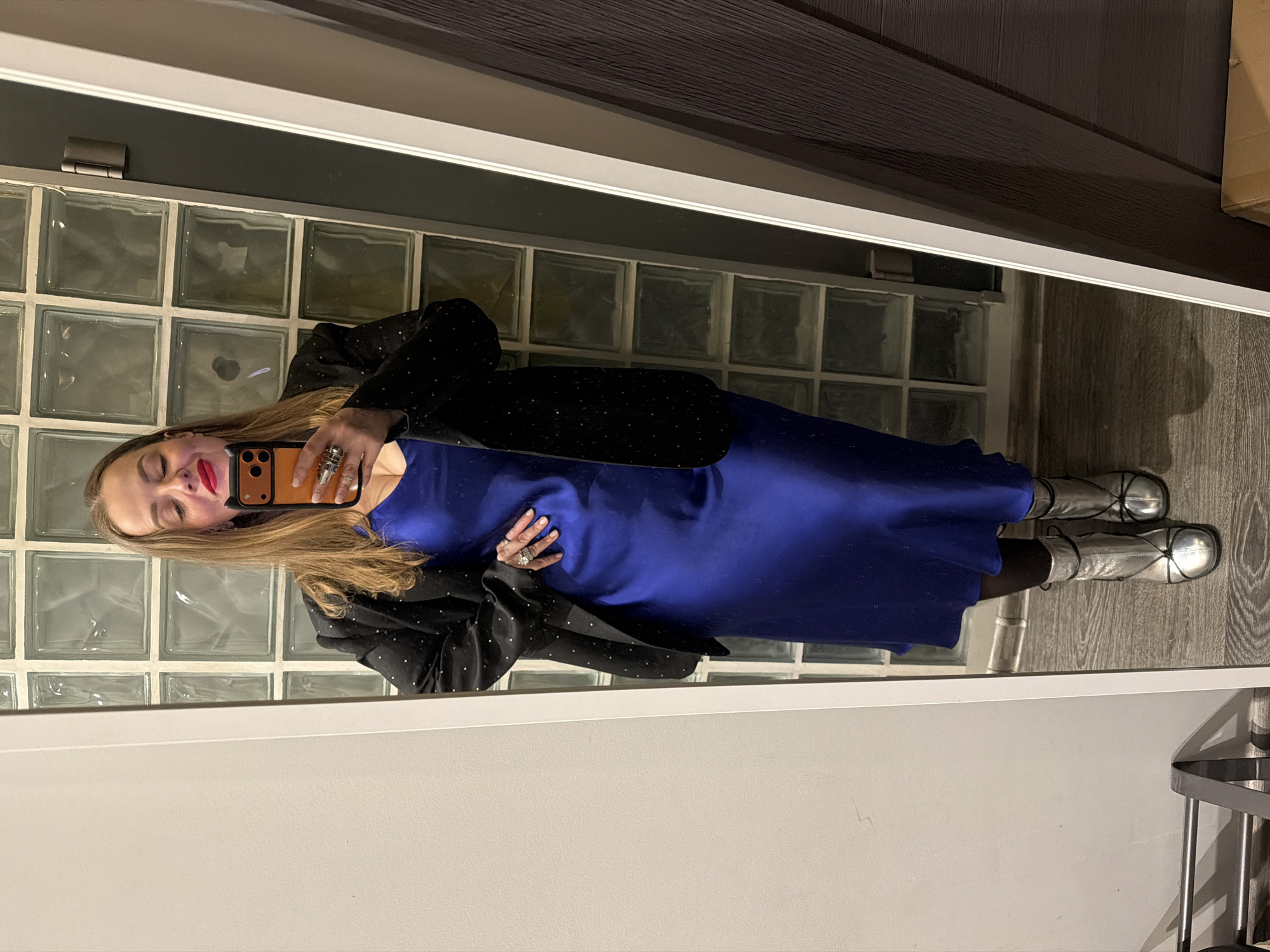 A person is standing in front of a mirror, taking a selfie. The mirror reflects a portion of the room.
Person:
- Gender: Female
- Attire: A blue dress with a black blazer with small white dots. She is also wearing silver boots.
- Accessories: Rings on her fingers.
- Hairstyle: Long and blond.
- Posture: Holding a smartphone to take a selfie. Her eyes are closed.
- Facial features: Red lipstick.
Background:
- Walls: A white or light-colored wall is visible in the foreground. The mirror reflects a wall with glass blocks. Above that is a dark-colored wooden-look wall.
- Floor: A light-colored wooden floor is visible.
Objects:
- Smartphone: Held by the person in the foreground.
- Mirror: Located in the foreground.
- Glass Blocks: In the background wall.
Moscow, Russia is not readily discernible from the visual elements in the image.