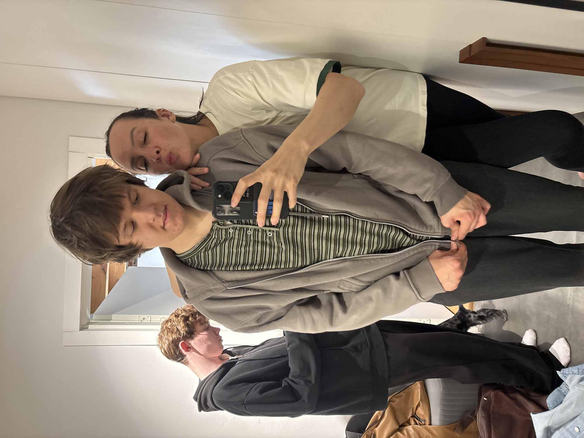 Three young individuals are captured in a vertical selfie indoors, facing a mirror. The person in the foreground, holding a dark-colored smartphone, has light brown hair and wears a grey zipped hoodie over a green and white striped long-sleeve shirt. Their left hand is adjusting the zipper of their hoodie, while their right hand holds the phone, directed towards themselves and the person behind them. Their eyes are mostly closed or looking down.
Behind them, a person with dark hair pulled back rests their head on the first person's shoulder, with eyes closed and lips pursed in a playful expression. They wear a cream-colored top with a dark green collar and black leggings. Their left arm is around the person in front. To the right of the selfie-taker, another individual with reddish-brown hair, wearing a black zipped hoodie, stands in profile, looking to their left (the viewer's right).
A small, fluffy dog with grey and black fur is visible between the legs of the two individuals on the right side of the frame. In the bottom right corner, a brown leather-like bag, a darker bag, and a folded light blue denim shirt are seen on the floor. The setting appears to be a room with light-colored walls, possibly white. A wooden shelf or beam is present on the wall in the upper right, and a window or doorway with a white frame is visible in the middle-left background, suggesting natural light. The overall mood is casual and affectionate. The location context is Chaikovsky, Russia. No visible text is clearly legible on any object.