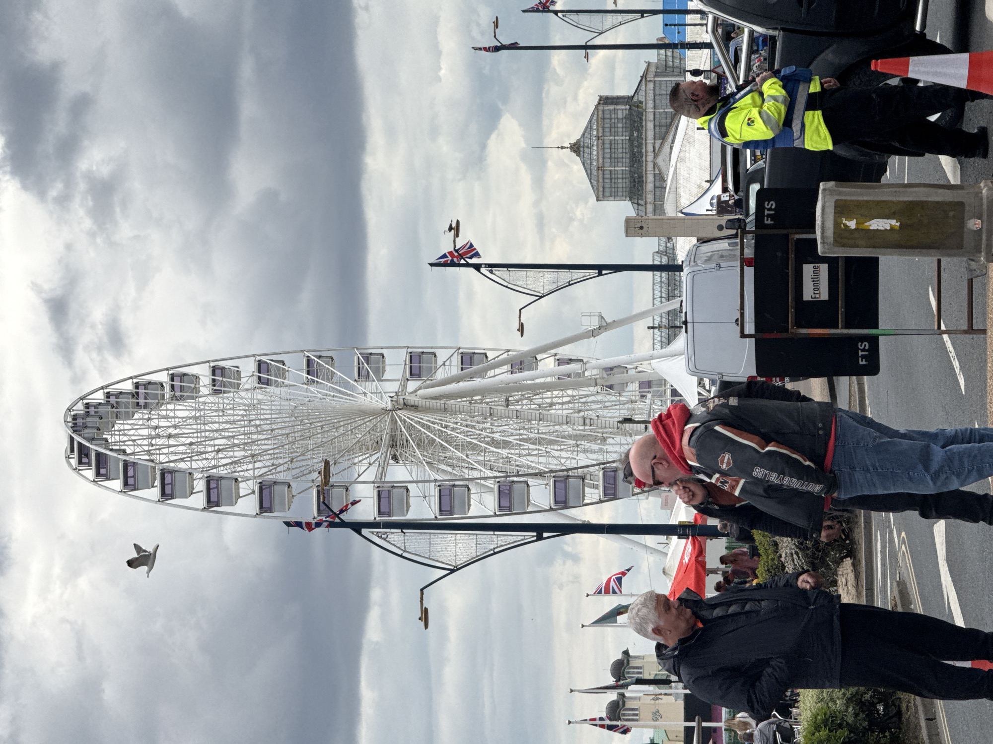 The photograph depicts a scene in Great Yarmouth, United Kingdom, dominated by the Ferris wheel, known as the Great Yarmouth Wheel. The sky is overcast. Union Jack flags are visible on lampposts and poles along the foreground. Several people are present on a paved area, including two men examining their fists, a man in a black jacket, and a safety guard in a yellow jacket leaning against a car. In the background, the Winter Gardens building is partially visible. A white van is parked behind the men, with a black sign marked "FTS" and "Frontline". A seagull is also visible in flight near the Ferris wheel.