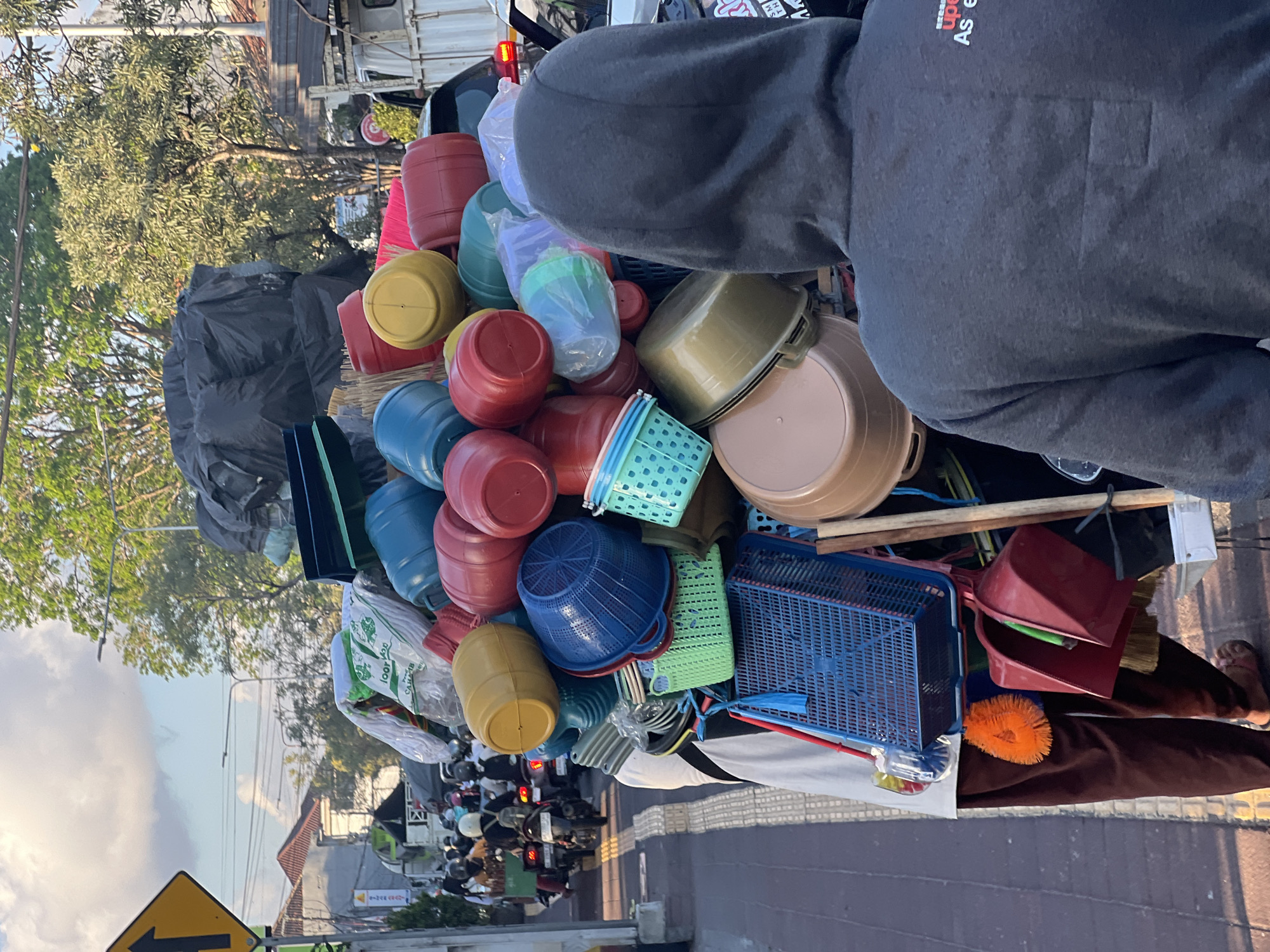 In Denpasar, Indonesia, a person wearing a dark grey hooded sweatshirt stands with their back to the camera, obscuring most of their face. They are positioned in the foreground, facing a heavily laden cart or vehicle. The cart is piled high with an assortment of plastic containers, including numerous red, blue, and yellow barrels, as well as various baskets and buckets in different colors and sizes. Some of the items are stacked precariously, and a few are wrapped in clear plastic. A green tarp is visible covering part of the load at the top.
To the left of the cart, a sidewalk with a patterned brick surface is visible, bordered by a white line. A yellow traffic sign with a black arrow pointing upwards is mounted on a pole on the far left. Beyond the sidewalk, a street is filled with traffic, primarily motorcycles, with riders wearing helmets. Buildings with tiled roofs line the street, and trees with green foliage are abundant, suggesting a tropical urban environment. The sky above is partly cloudy. The person in the foreground appears to be either assisting with or observing the transportation of these goods. The overall scene depicts a busy street in Denpasar, with a focus on the individual and the extensive collection of plastic items.