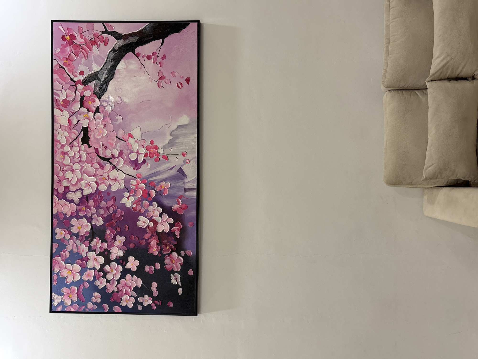 The image displays a painting of pink cherry blossoms on a dark background with a greyish-pink sky. The painting is rectangular and framed in black. It is hung on a light-colored wall. Below the painting, a portion of a beige sofa with cushions is visible. The scene appears to be an indoor setting, likely a living room, within a residential property in Binong, Indonesia. There are no people visible in the image.