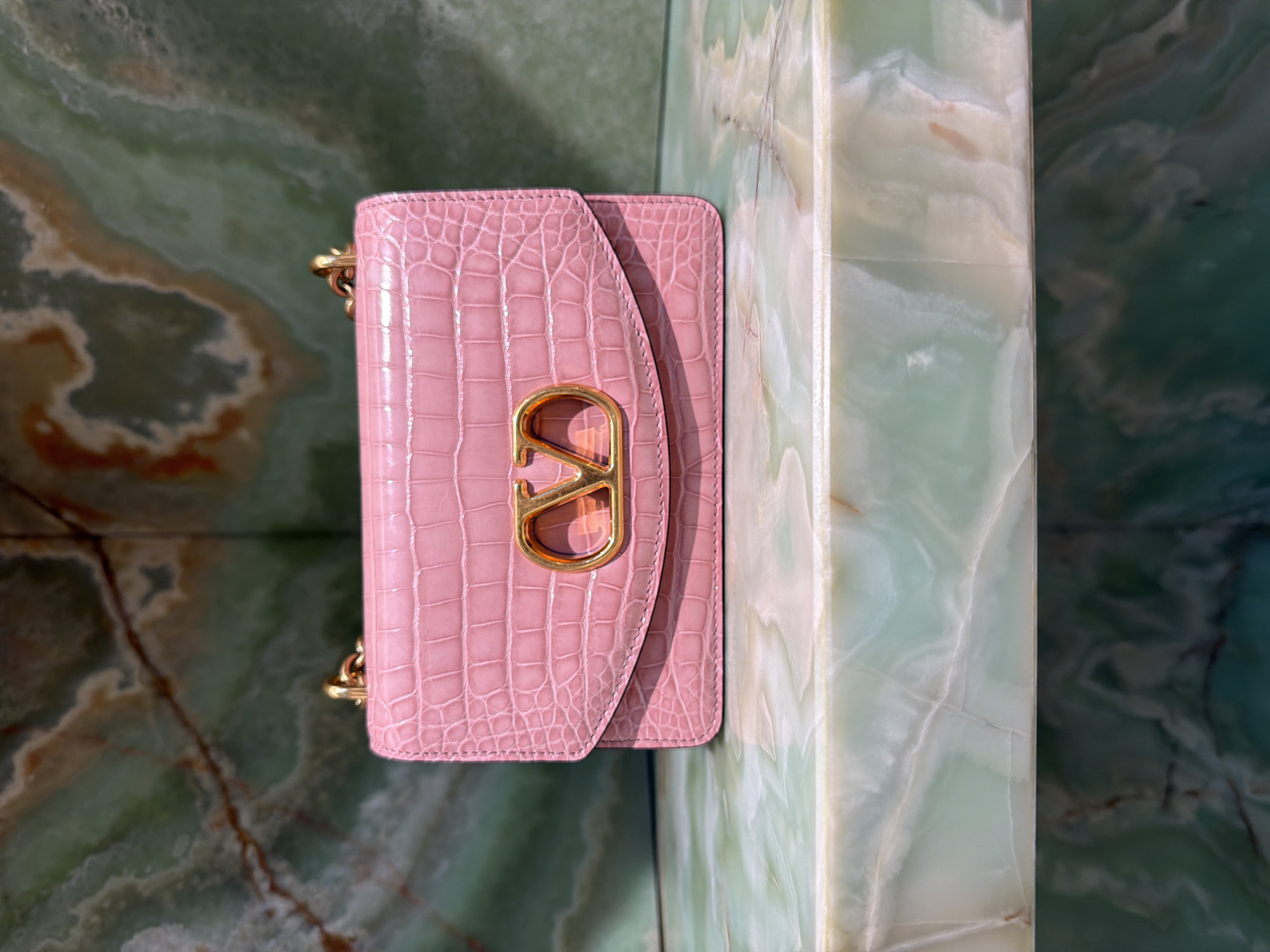 A pink, crocodile-embossed Valentino handbag with a gold V-logo is displayed on a polished, pale green marble surface. The handbag has a flap closure and a gold chain strap. The background consists of a wall with a green, marble-like pattern. The scene is indoors, likely a retail or display setting. The image does not contain any people or actions.