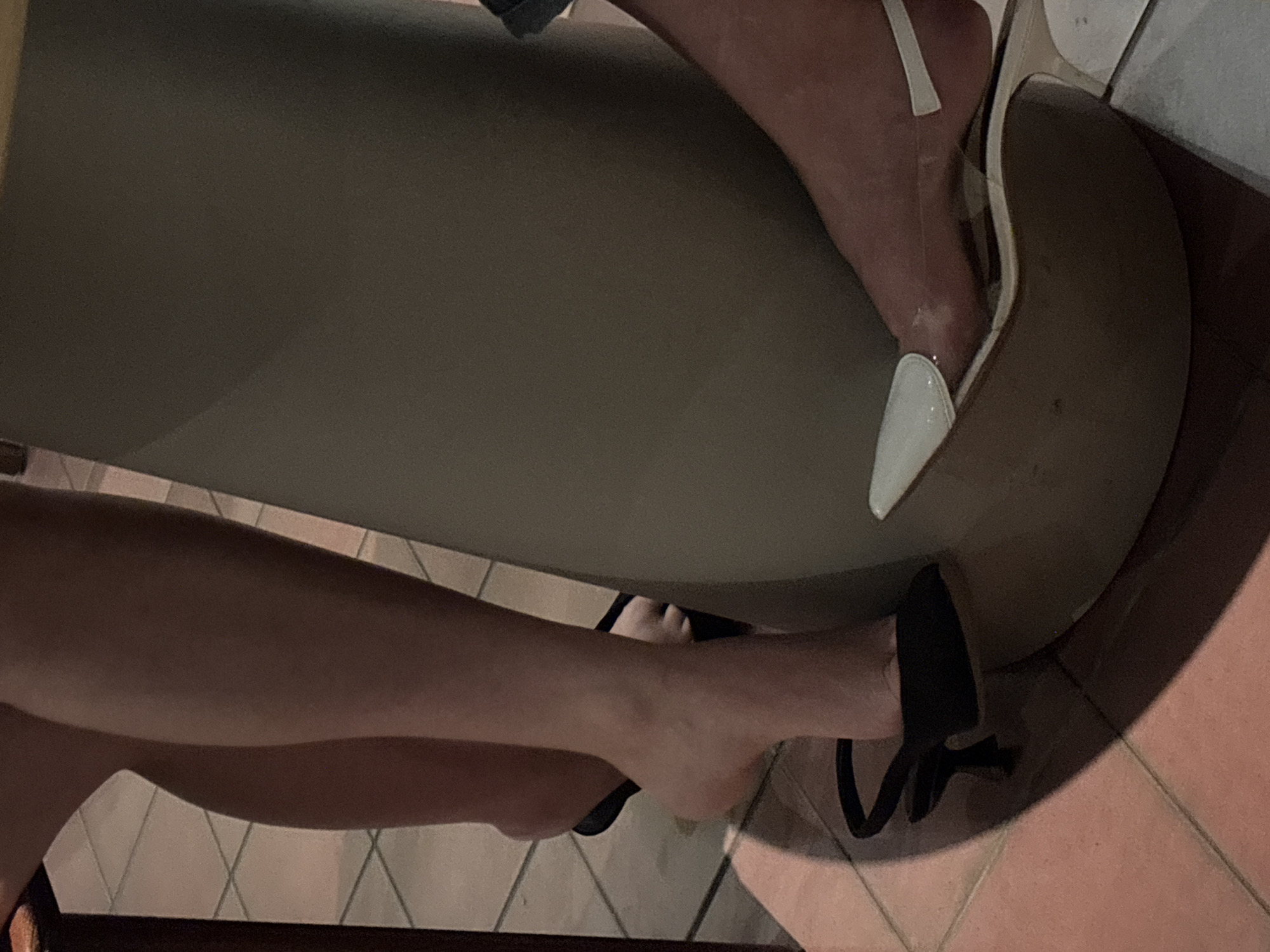 The image shows a close-up view of two people's legs and feet. One person is wearing black heels, and the other is wearing clear heels. Both are standing near a large, cylindrical object, possibly a table or a pillar, which is light brown in color. The floor is tiled with a pinkish-brown hue. The lighting suggests an indoor setting, possibly a restaurant or bar. The image does not contain any information that would allow for determining its location in Novaya Usman', Russia.