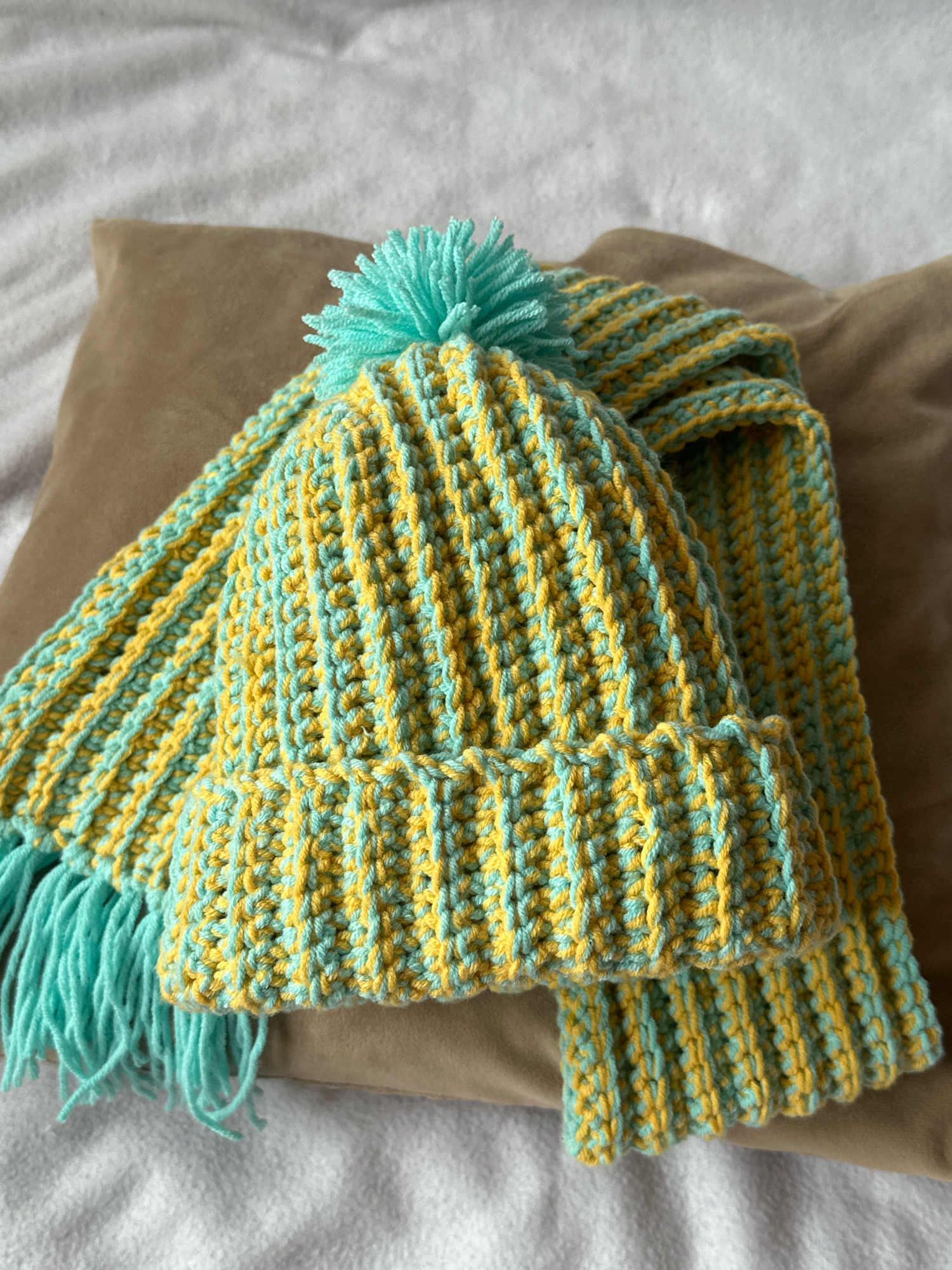 A knitted hat and scarf set, in alternating bands of yellow and turquoise, rests on a brown pillow. The hat has a turquoise pom-pom on top. The scarf has turquoise fringe. The background is out of focus, showing a textured white surface and the brown pillow. There are no people or actions depicted. The scene is indoors.