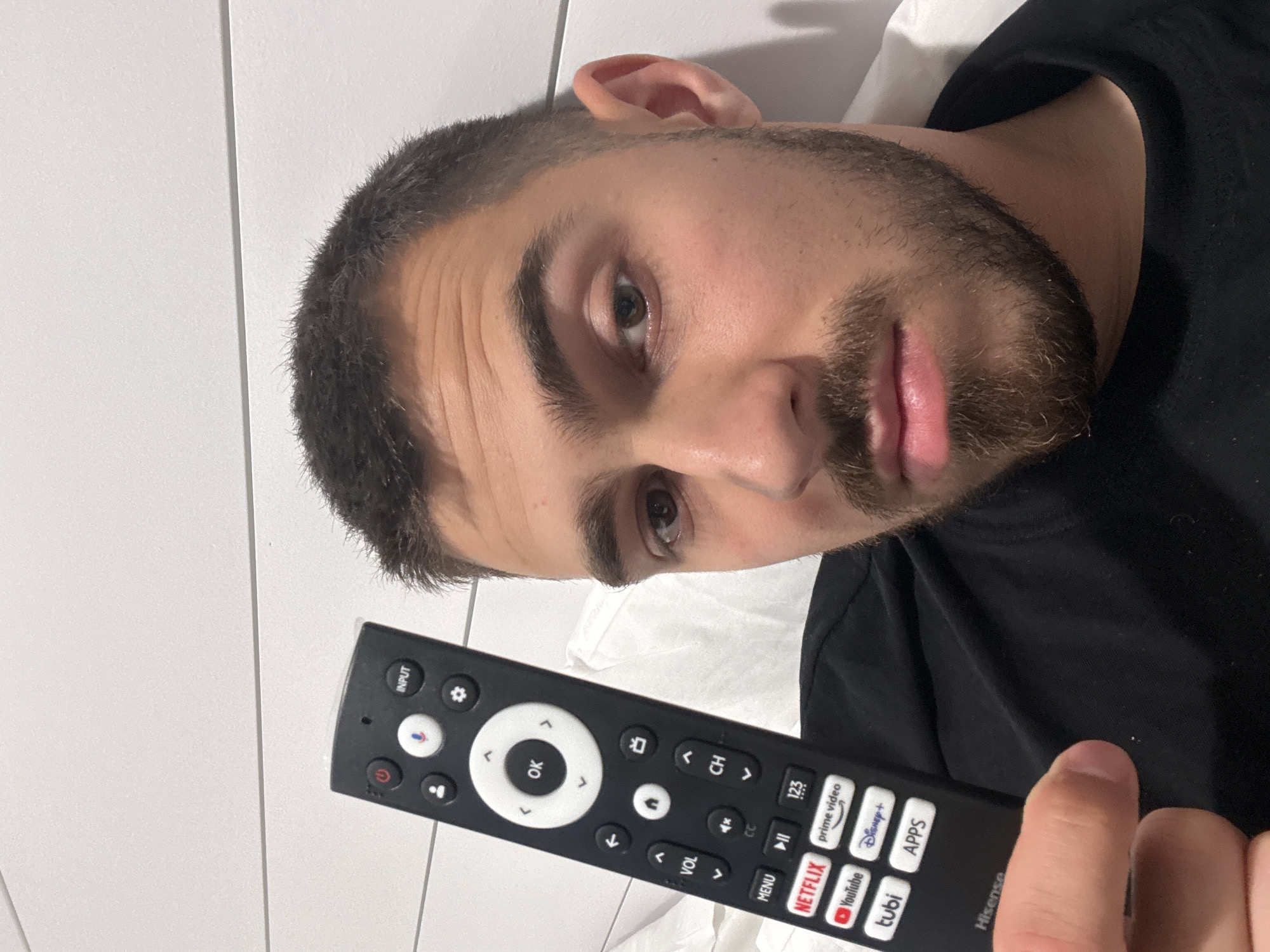 A man is holding a black Hisense TV remote control in his right hand. The remote has buttons for power, input, settings, volume, channel, menu, and quick access to streaming apps such as Netflix, YouTube, Disney+, Prime Video, and Tubi. The man has short, dark hair and a beard. He is wearing a black shirt and lying on a white pillow. The background is a white wall with horizontal lines. The image was likely taken indoors. The location is impossible to determine.