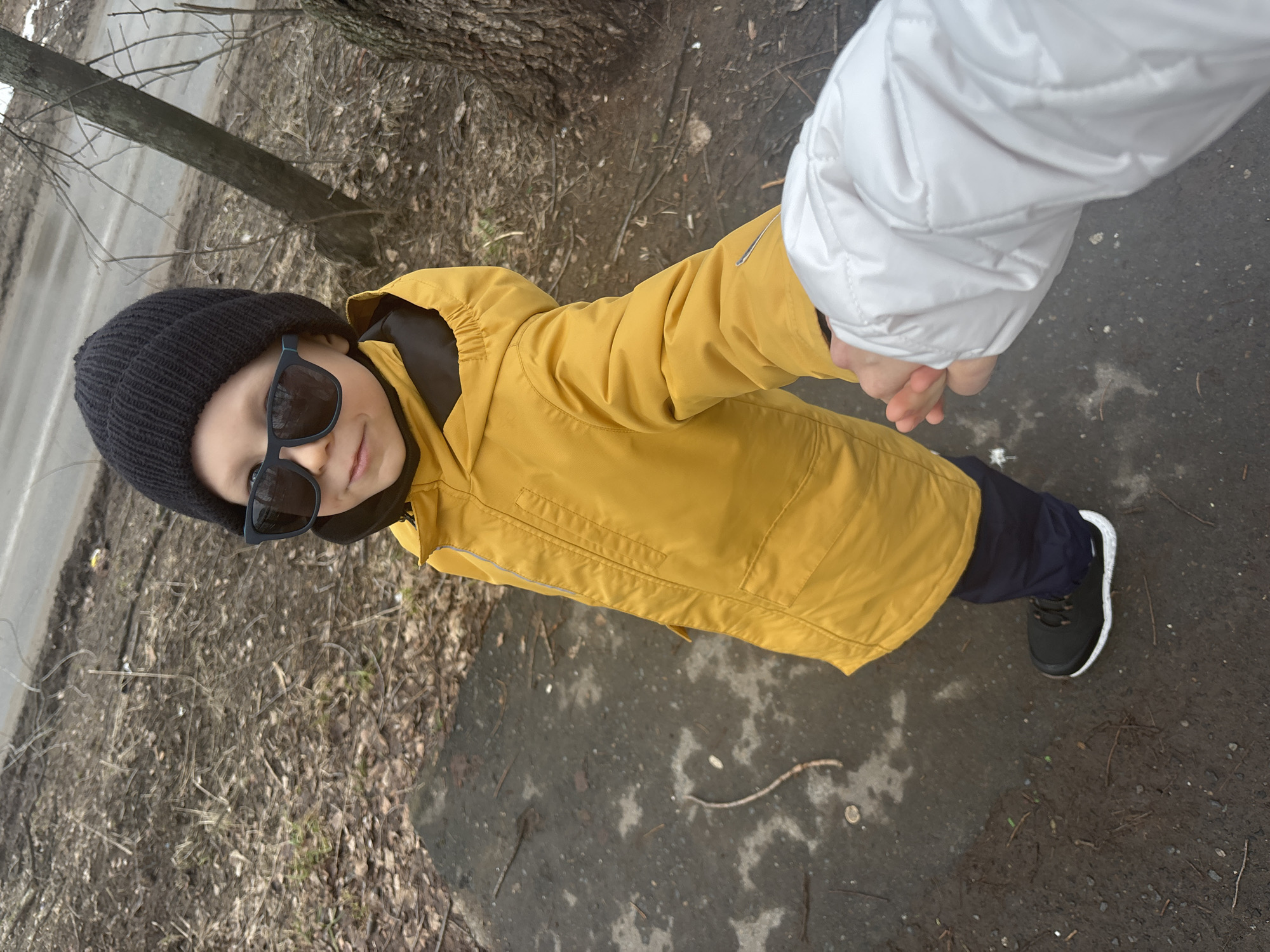 A child wearing a black beanie, sunglasses, a yellow jacket, and black pants with black and white sneakers stands on a weathered concrete surface adjacent to a patch of dry, brown vegetation. The child is holding hands with a person wearing a white puffy jacket. A tree trunk and a bare tree branch are visible to the left of the child. A road can be seen in the background. The image was taken in Chaikovsky, Russia.