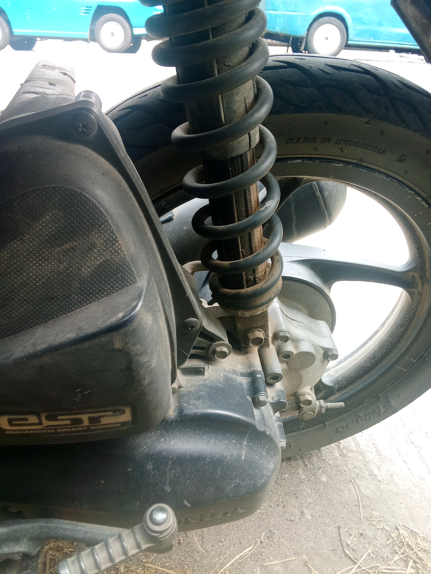 The image shows a close-up view of the rear shock absorber and tire of a scooter. The scooter is parked on a dirt or gravel surface, possibly a road or driveway, with dry grass and twigs scattered around. In the background, a portion of a blue vehicle, possibly a car or van, is visible. The scooter's tire has "MADE IN INDONESIA" embossed on its sidewall. The scooter also has a black body panel with the text "eSP ENHANCED SMART POWER" written on it in gold lettering. A kick starter lever is visible in the lower part of the frame. The overall scene appears to be outdoors in an urban or semi-urban environment. There is no information in the image to determine its specific location within Selaraja, Indonesia, beyond the country of origin indicated on the tire.