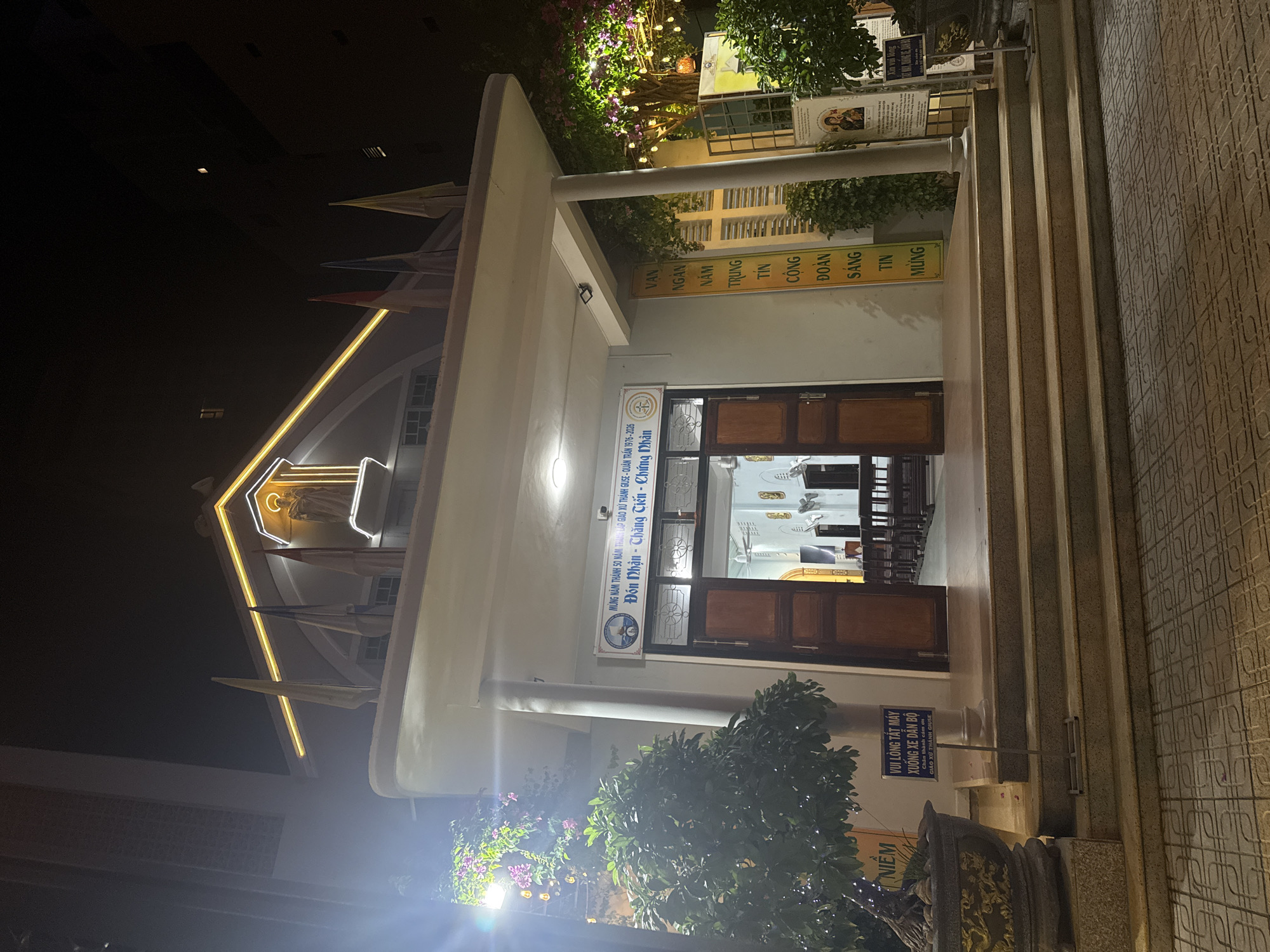 The image shows the exterior of the Giáo Xứ Thánh Giuse Church (Saint Joseph Parish) in Nha Trang Ward, Vietnam, at night. The church facade is illuminated with a light at the top, along with some white flags. At the front of the building, there's a small sign indicating the parish was established in 1976 and will be celebrating 50 years in 2026. A few plants, including a small bush with flowers, are near the entrance of the church. A sign reads, "Please turn off engine. Dismount motorbike. Thank you for your cooperation." The steps leading to the entrance are also visible, and a sign on the wall on the right lists the words "Vạn Ngàn Năm", "Trung Tín", "Cộng Đoàn" and "Sáng Tin Mừng".